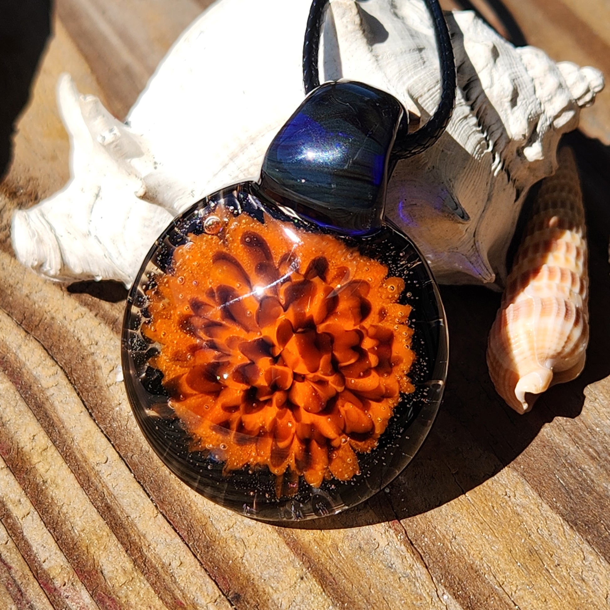 Fiery Glass Pendant Necklace resting on seashells, showcasing its bright orange bloom and glossy black glass. Handmade Halloween accessory.