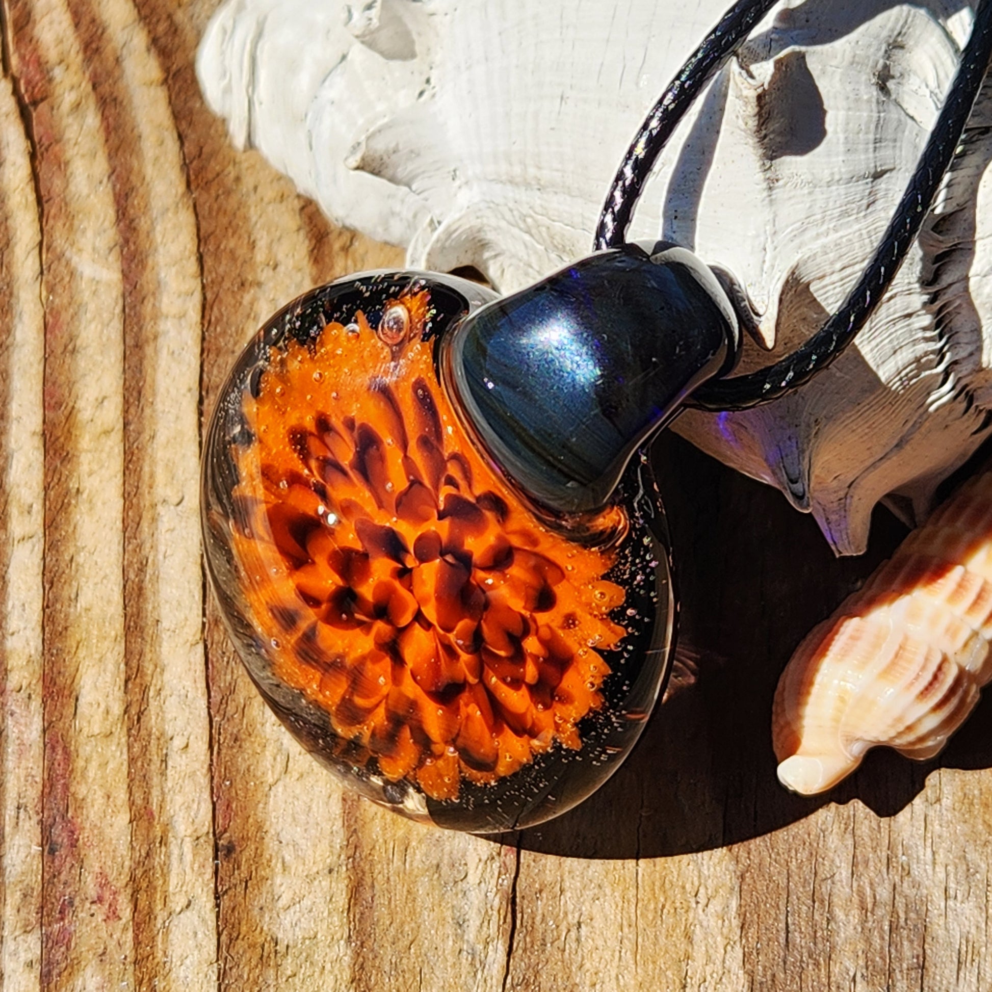 Fiery Flame Glass Pendant on wood background with seashells, highlighting its vivid orange bloom and glossy black glass. Handmade fall necklace.