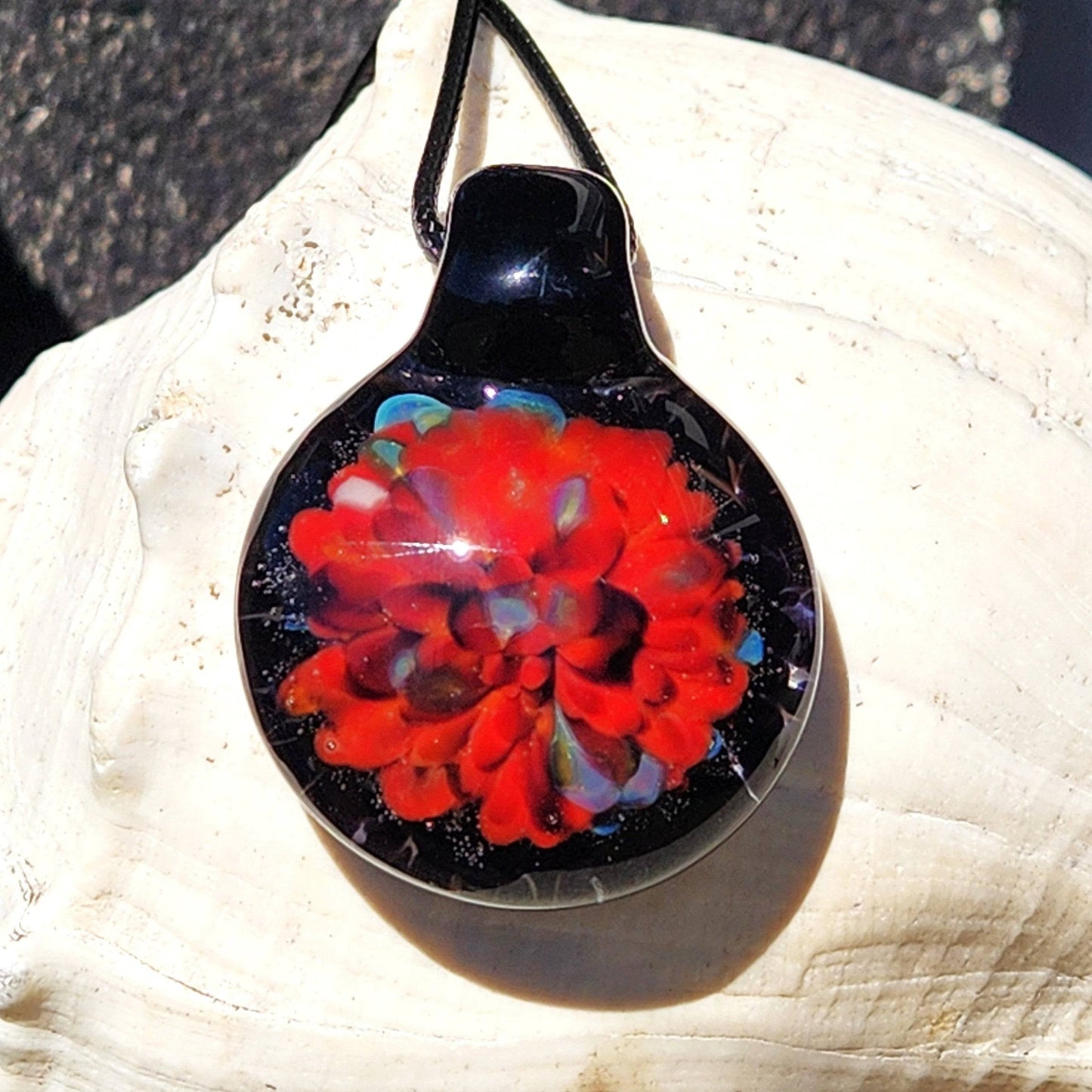 One-of-a-kind red floral glass pendant necklace resting on a white shell. Handmade borosilicate jewelry with vivid color and durable craftsmanship.