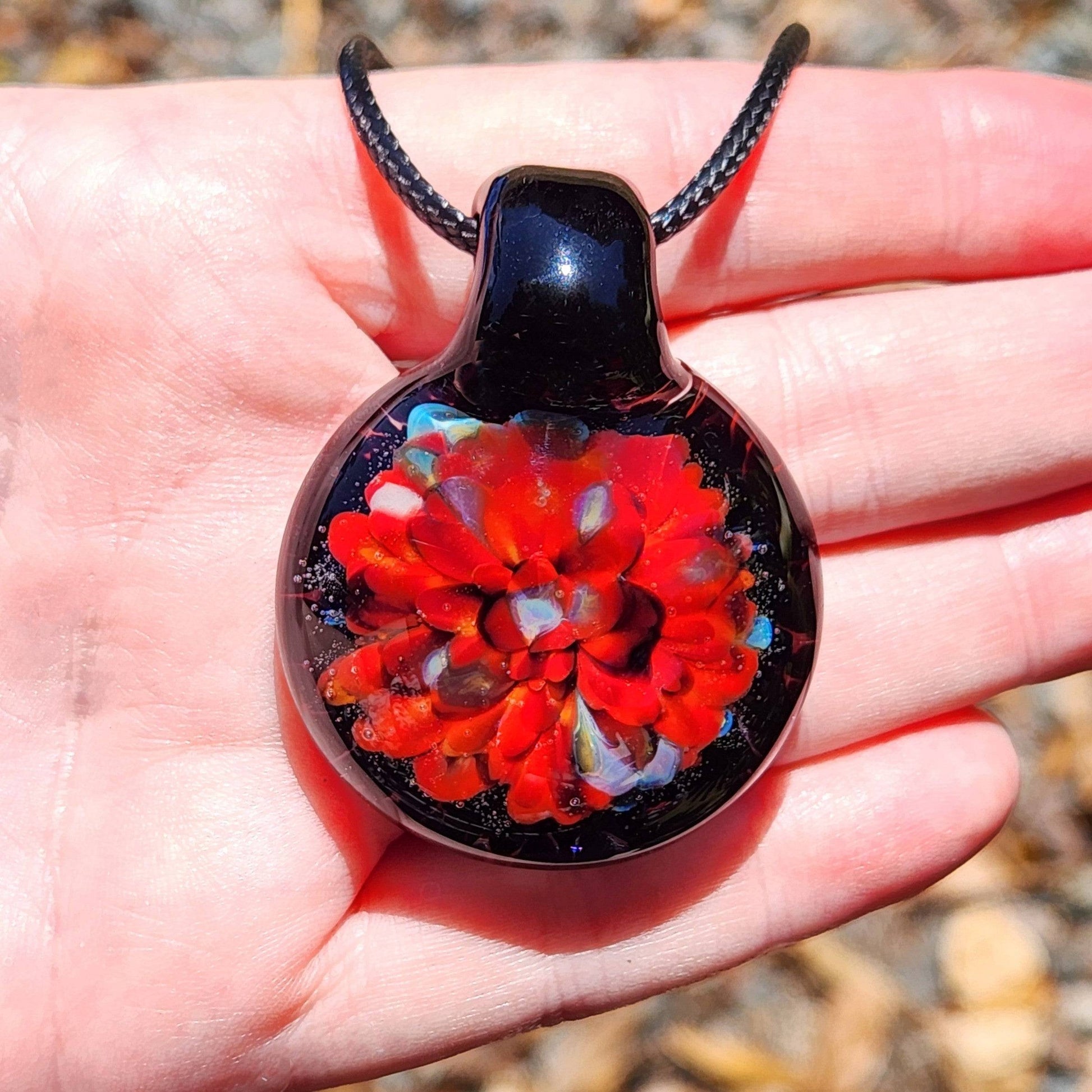 Radiant red glass pendant necklace displayed in hand for size reference. Handmade blown glass jewelry with vivid floral design and black base.