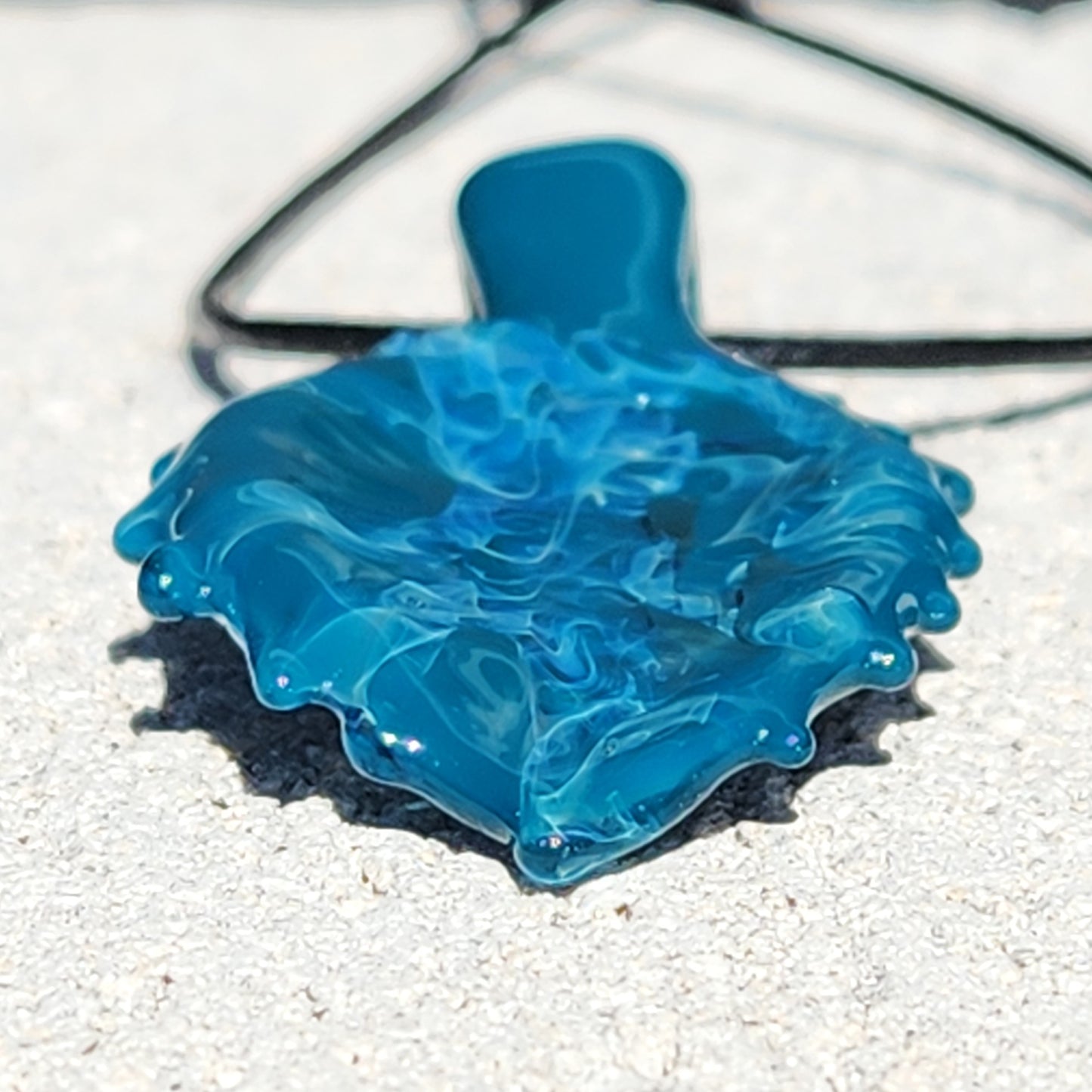 Close-up of Ocean Drift Glass Pendant Necklace on sand, showcasing its rippled glass surface and vivid oceanic color blend.