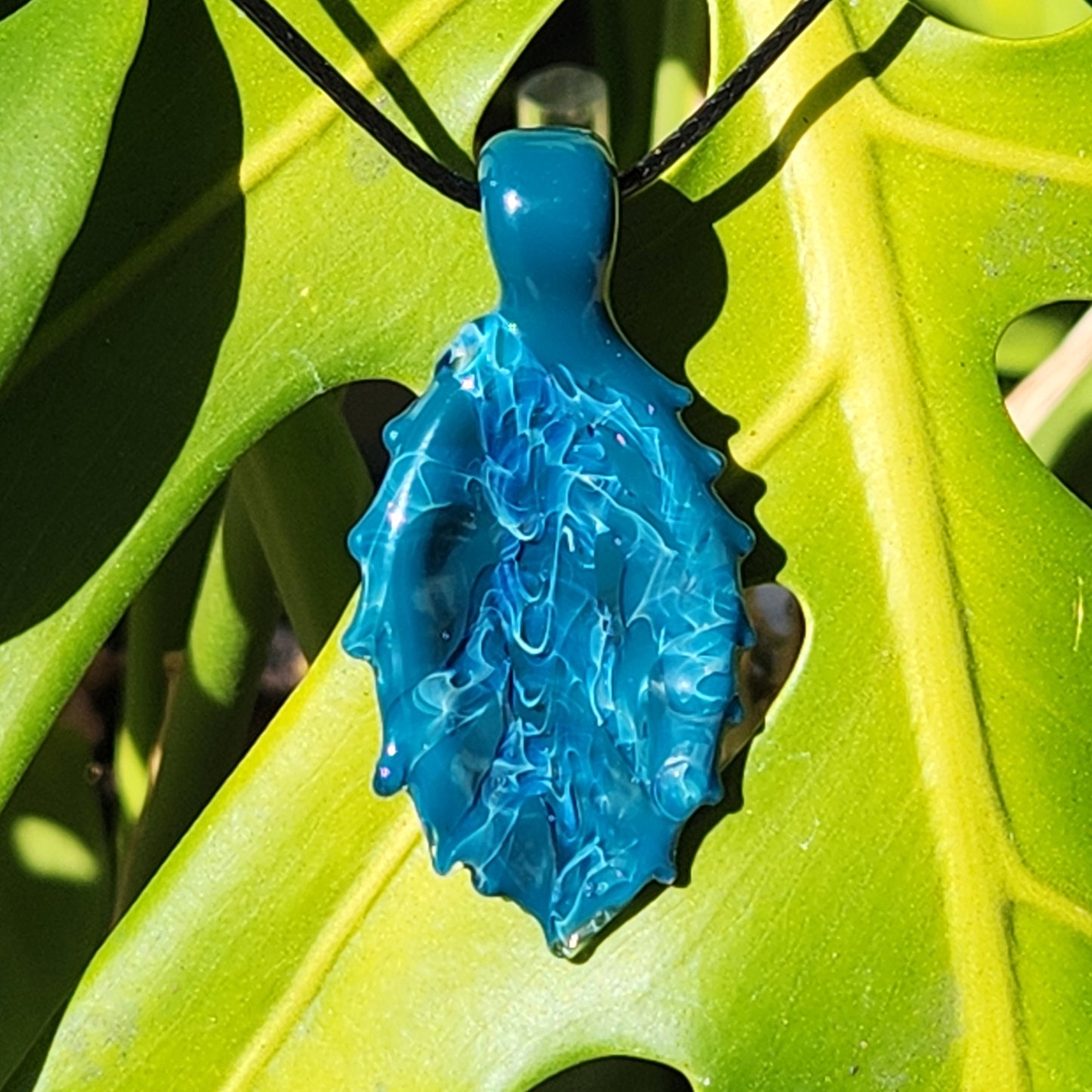 Ocean Drift Glass Pendant Necklace hanging on a green leaf, capturing its vibrant blue-green color shift in natural light.