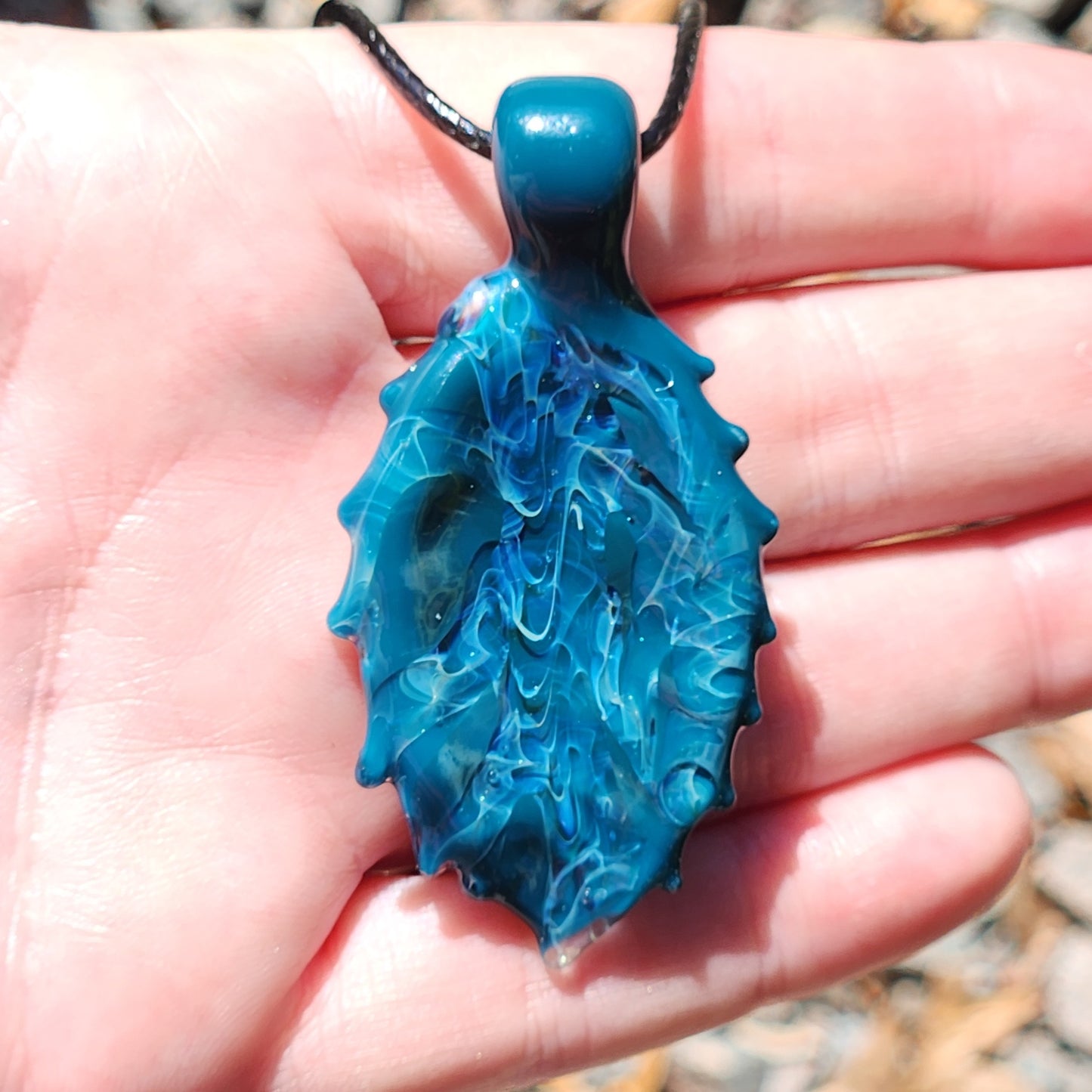 Front view of Ocean Drift Glass Pendant Necklace in hand, highlighting its swirling blue glass and handmade silver-infused details.