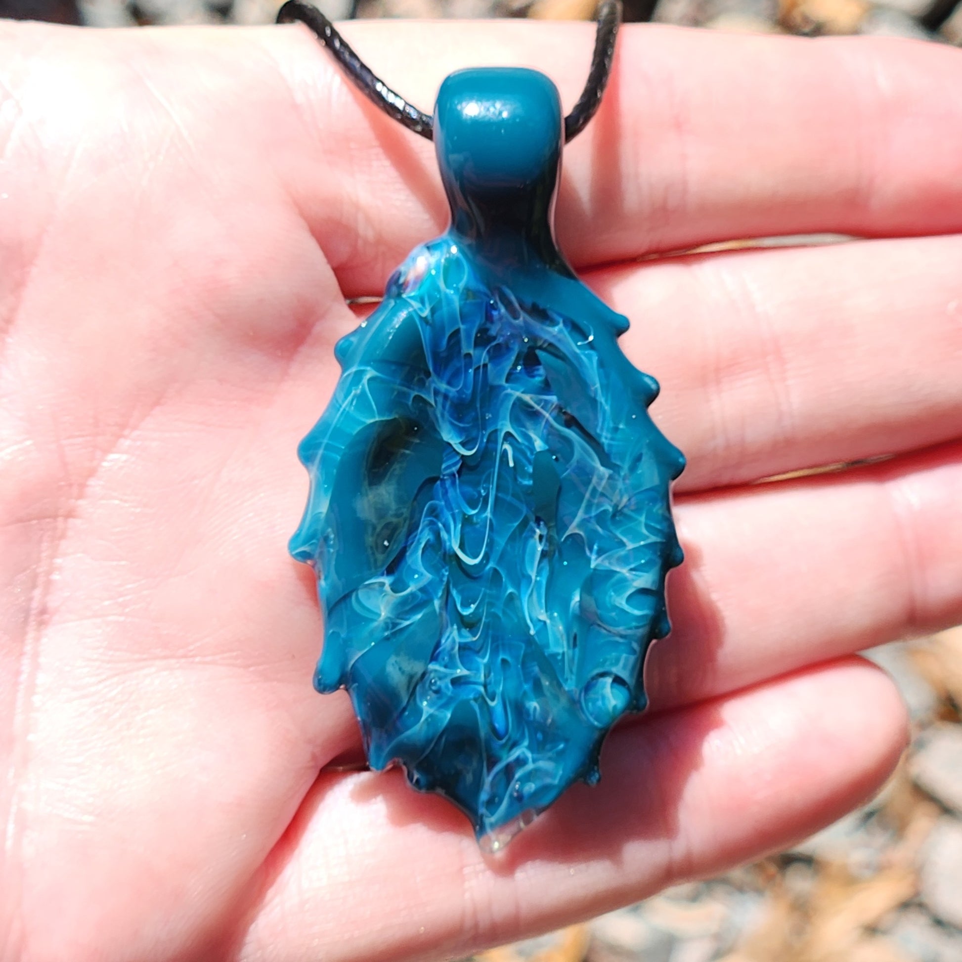 Front view of Ocean Drift Glass Pendant Necklace in hand, highlighting its swirling blue glass and handmade silver-infused details.