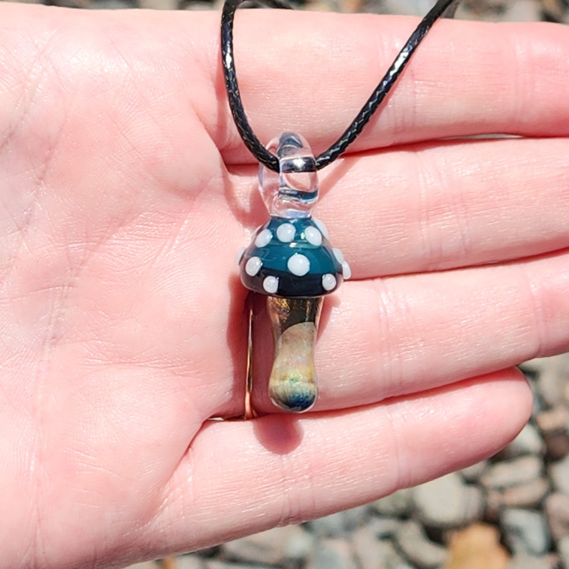 Hand holding Blue Mushroom Art Glass Pendant Necklace with metallic stem and blue cap. A unique handmade accessory for nature enthusiasts.