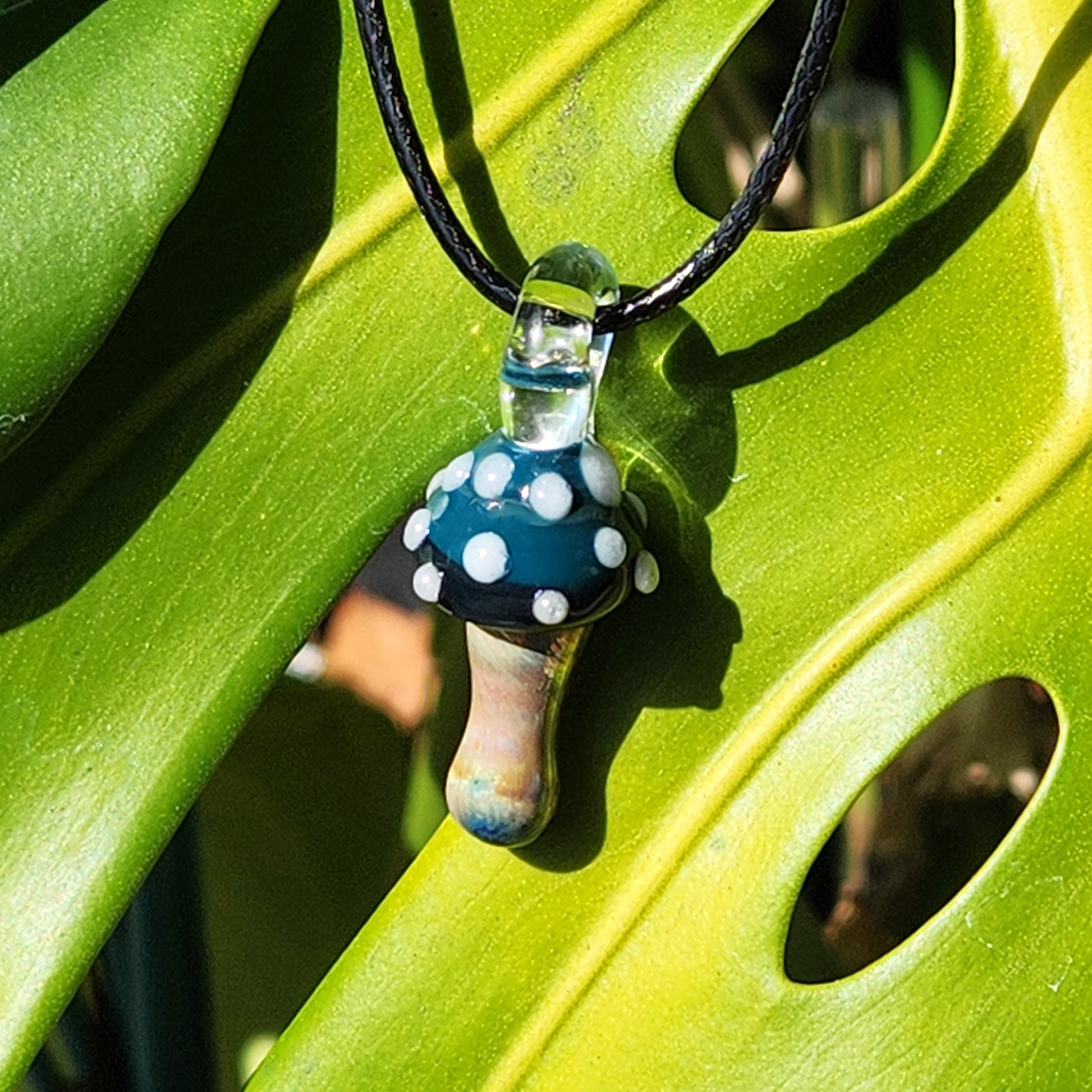 Blue Mushroom Pendant Necklace hanging on a green leaf, highlighting its natural theme and boho aesthetic. Handmade glass jewelry piece.