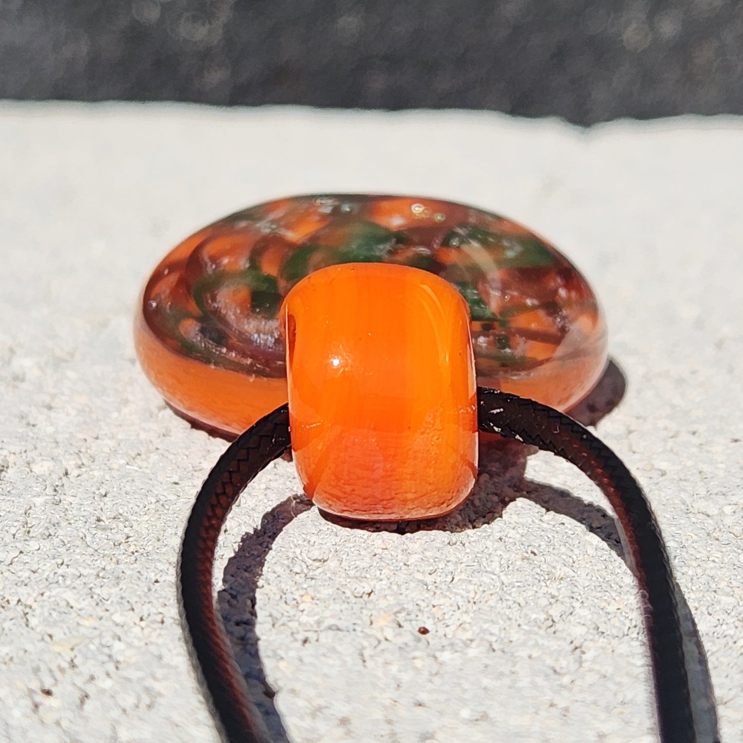 Close-up of the orange glass loop on the pendant necklace, showing smooth craftsmanship and black cord threading.