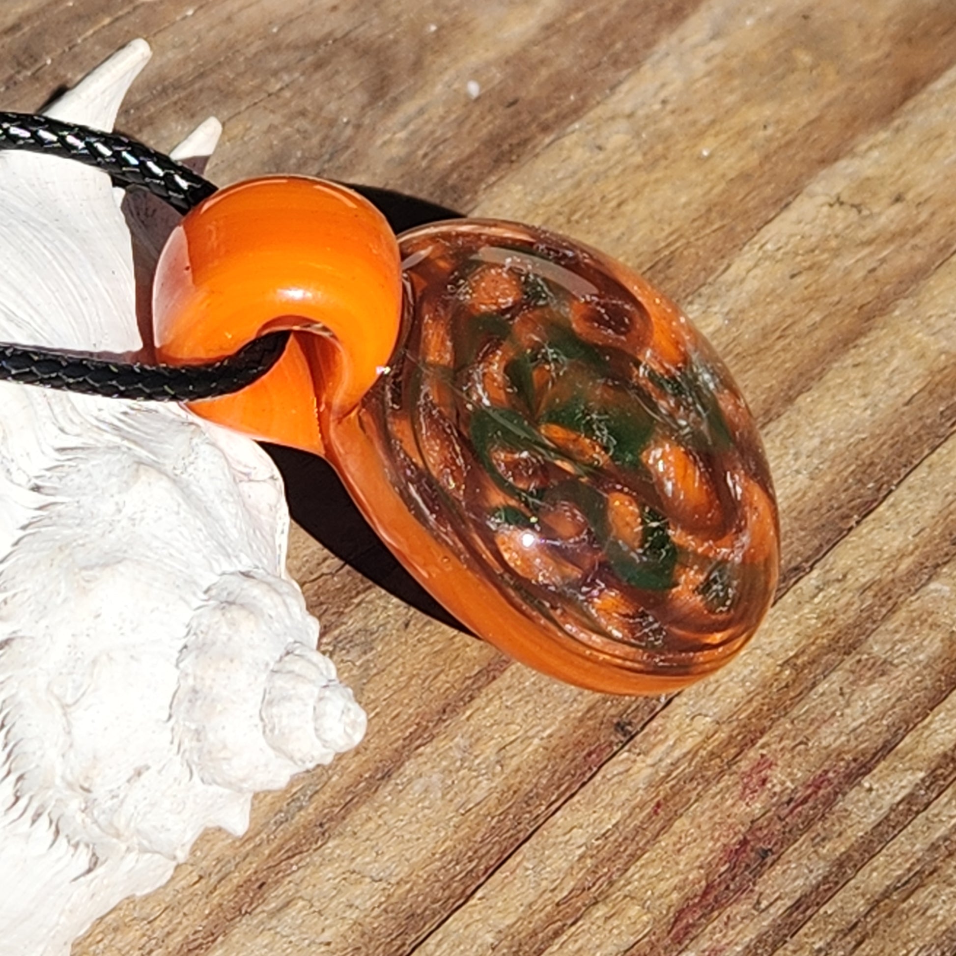 Angled view of handmade orange glass pendant with green and silver swirls, resting on a natural wood surface.