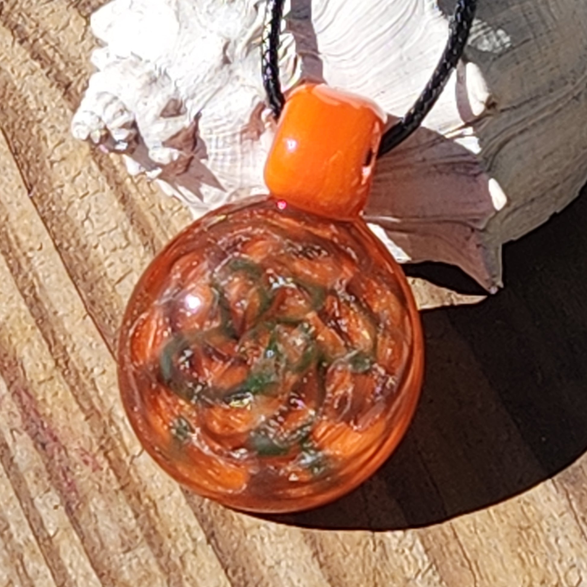 Handmade orange glass pendant necklace resting on wood, featuring swirling green and silver sparkles in a boho style.