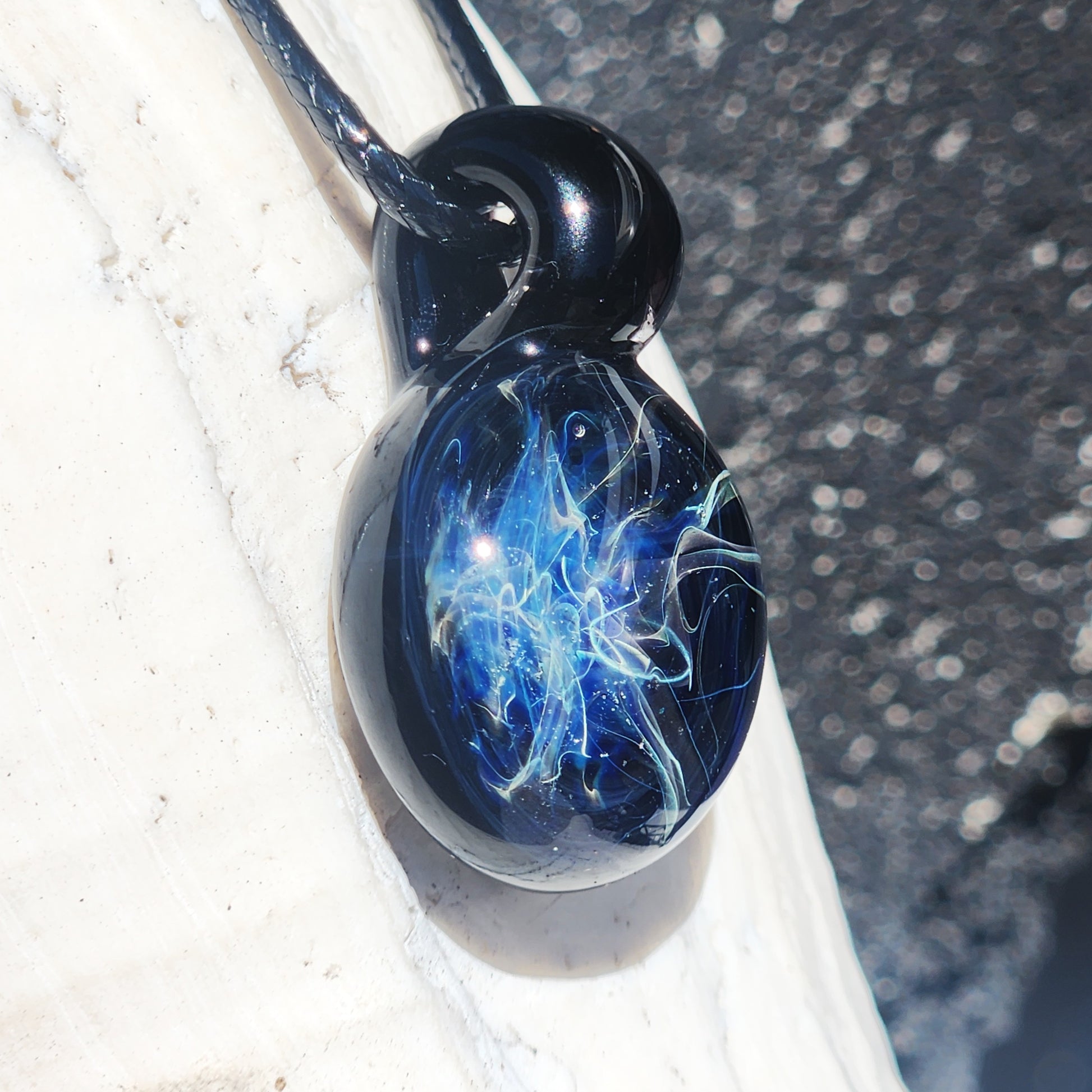 Artisan glass pendant with glowing blue and silver swirls, photographed on a white shell to highlight its handcrafted detail.