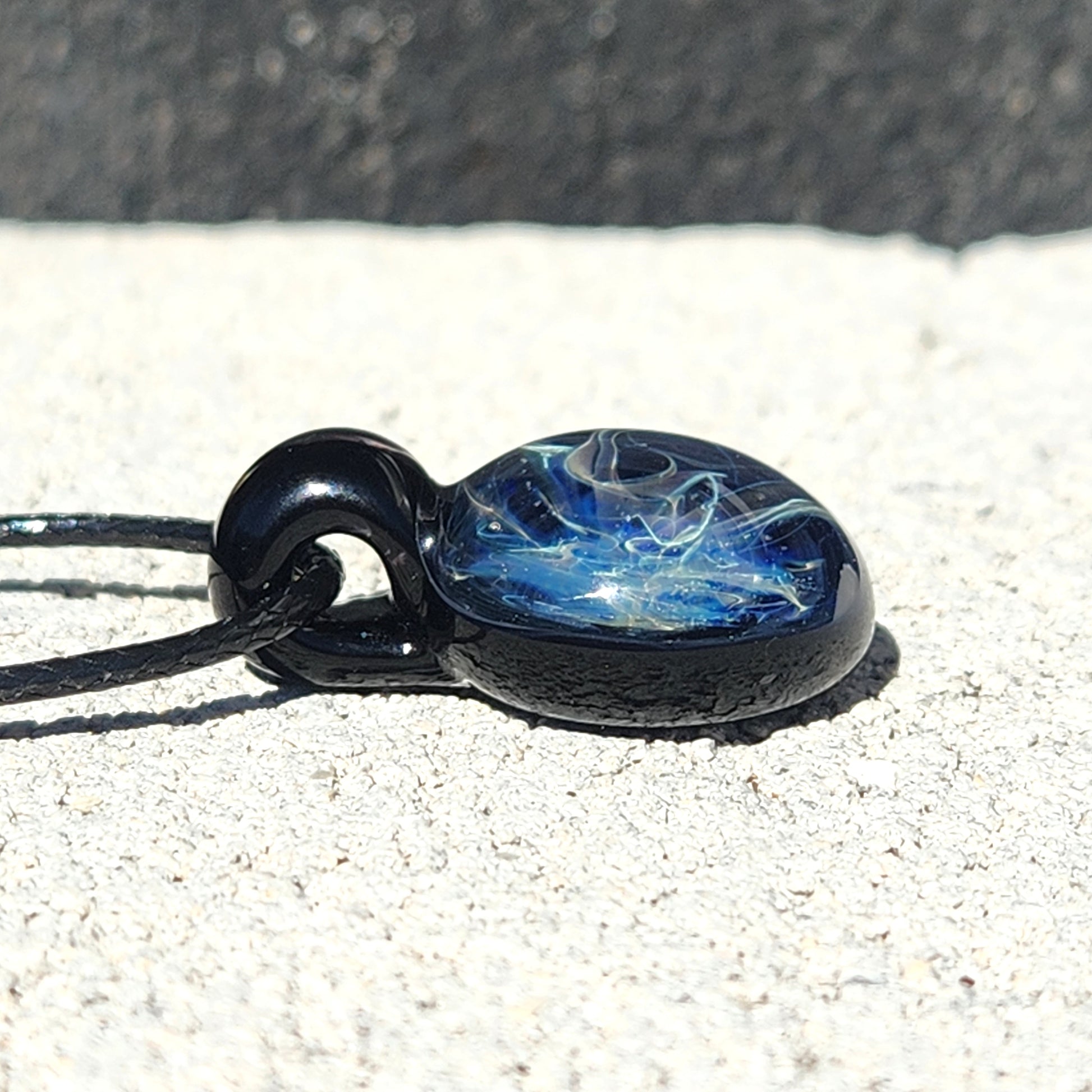 Low-angle view of small handmade glass pendant necklace with deep blue and silver galaxy-inspired design on black cord.