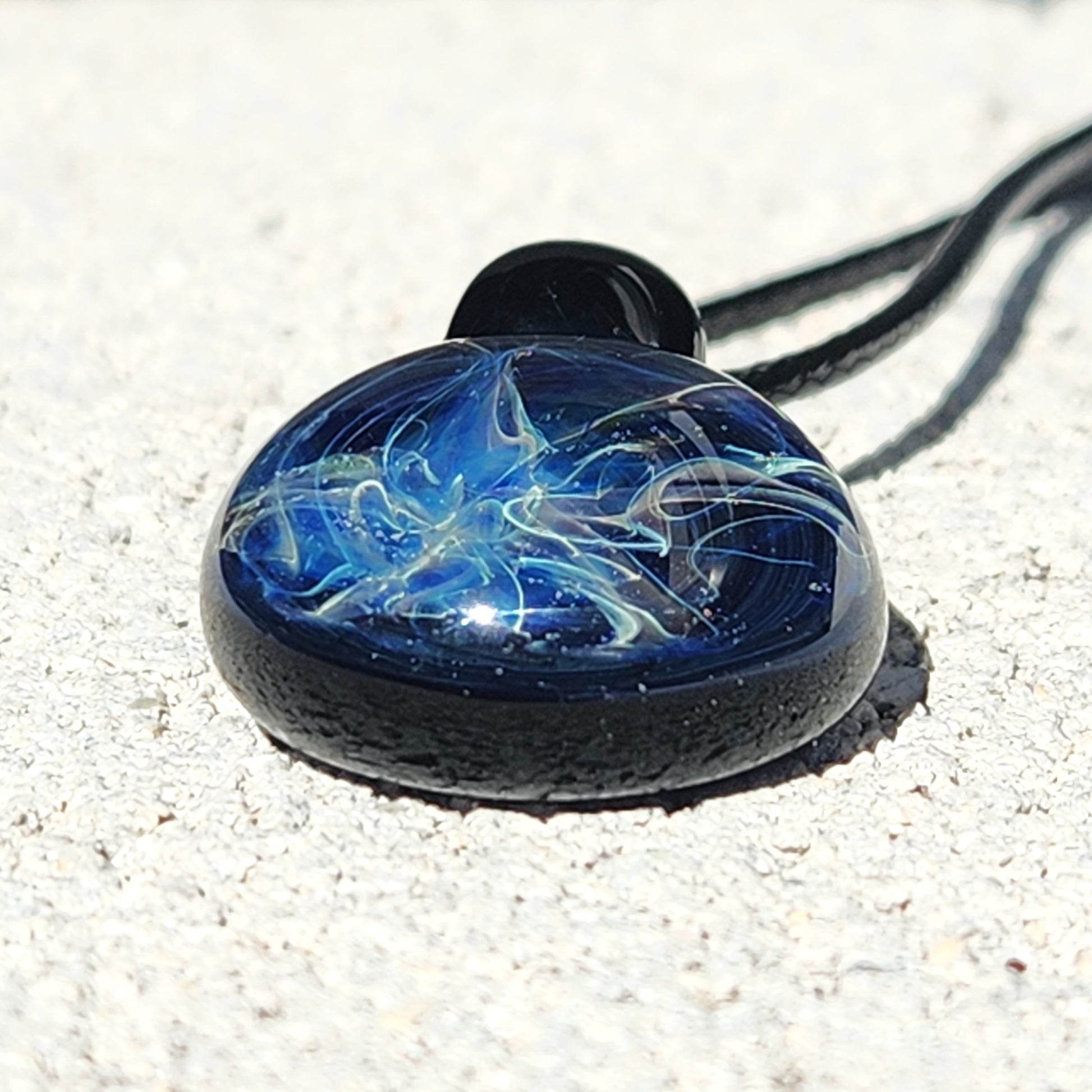 Close-up of handmade glass pendant necklace with galaxy-like blue and silver swirls, resting on a light stone surface.