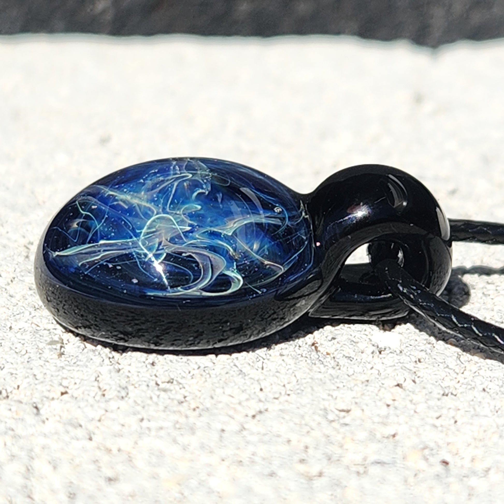 Side view of handmade blown glass pendant necklace with black loop and vivid blue swirl pattern, lying on textured stone.