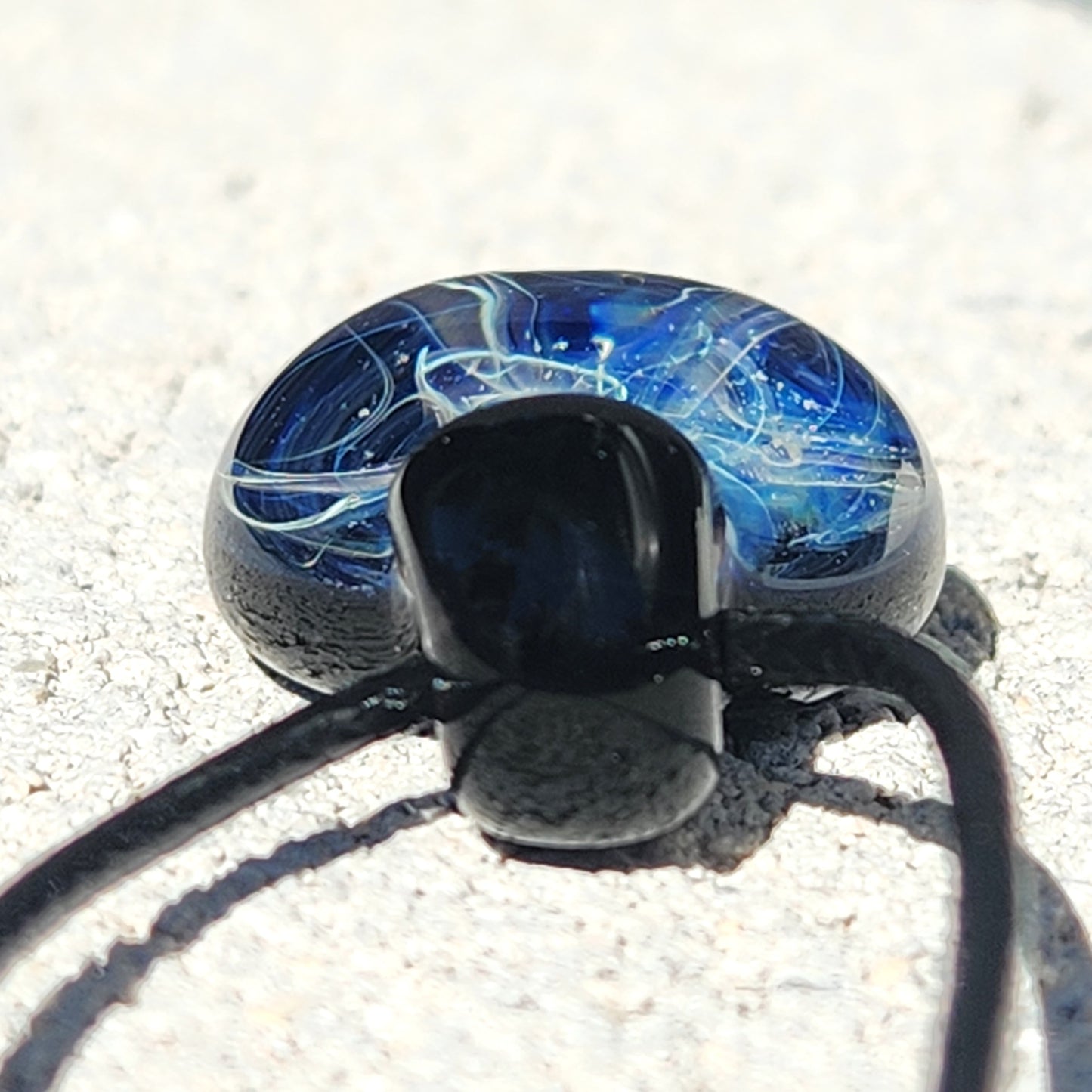 Bottom view of handmade glass pendant necklace showing detailed swirl design and black glass loop on a sandy surface.