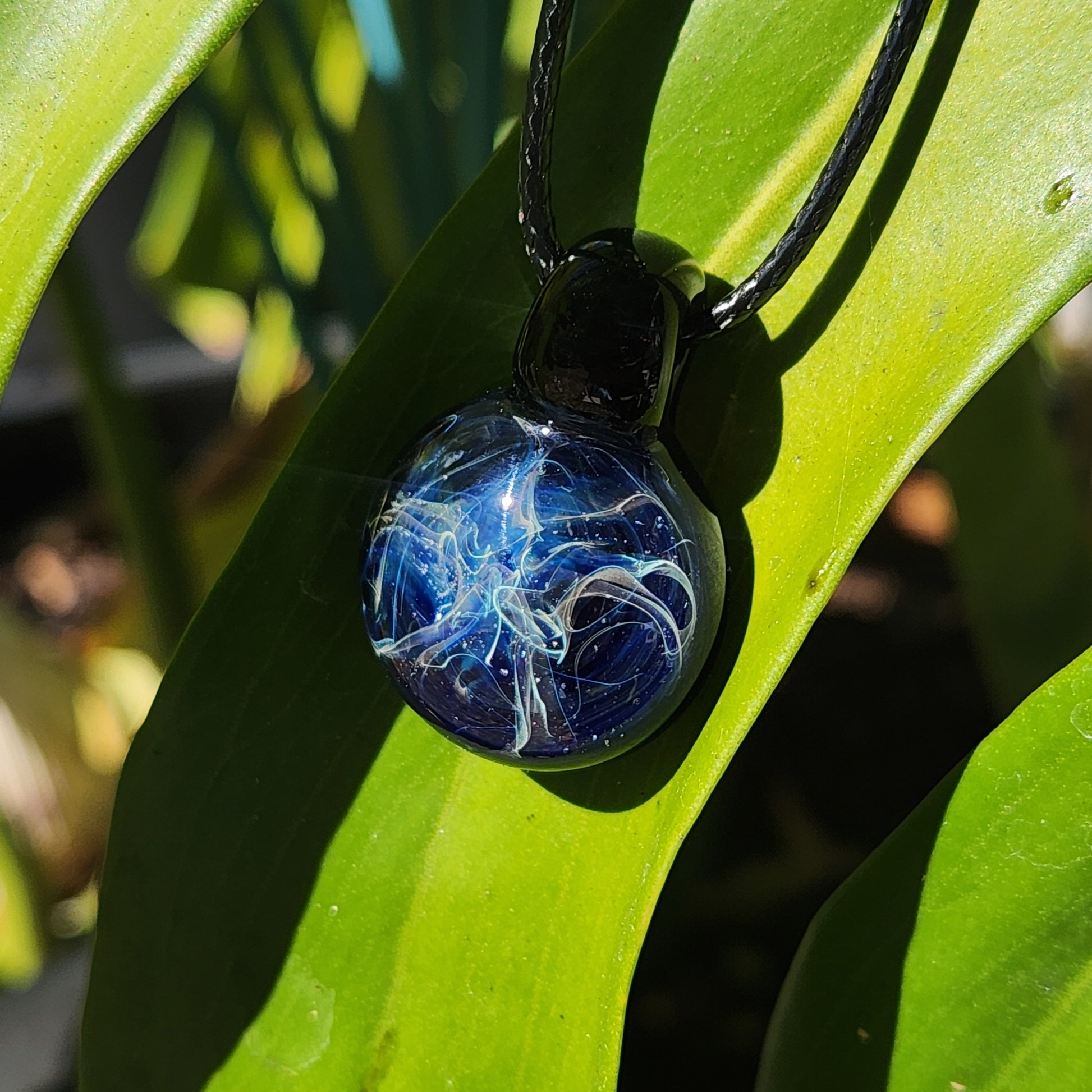 Handblown glass pendant necklace with swirling blue and silver colors, hanging against a vibrant green leaf background.