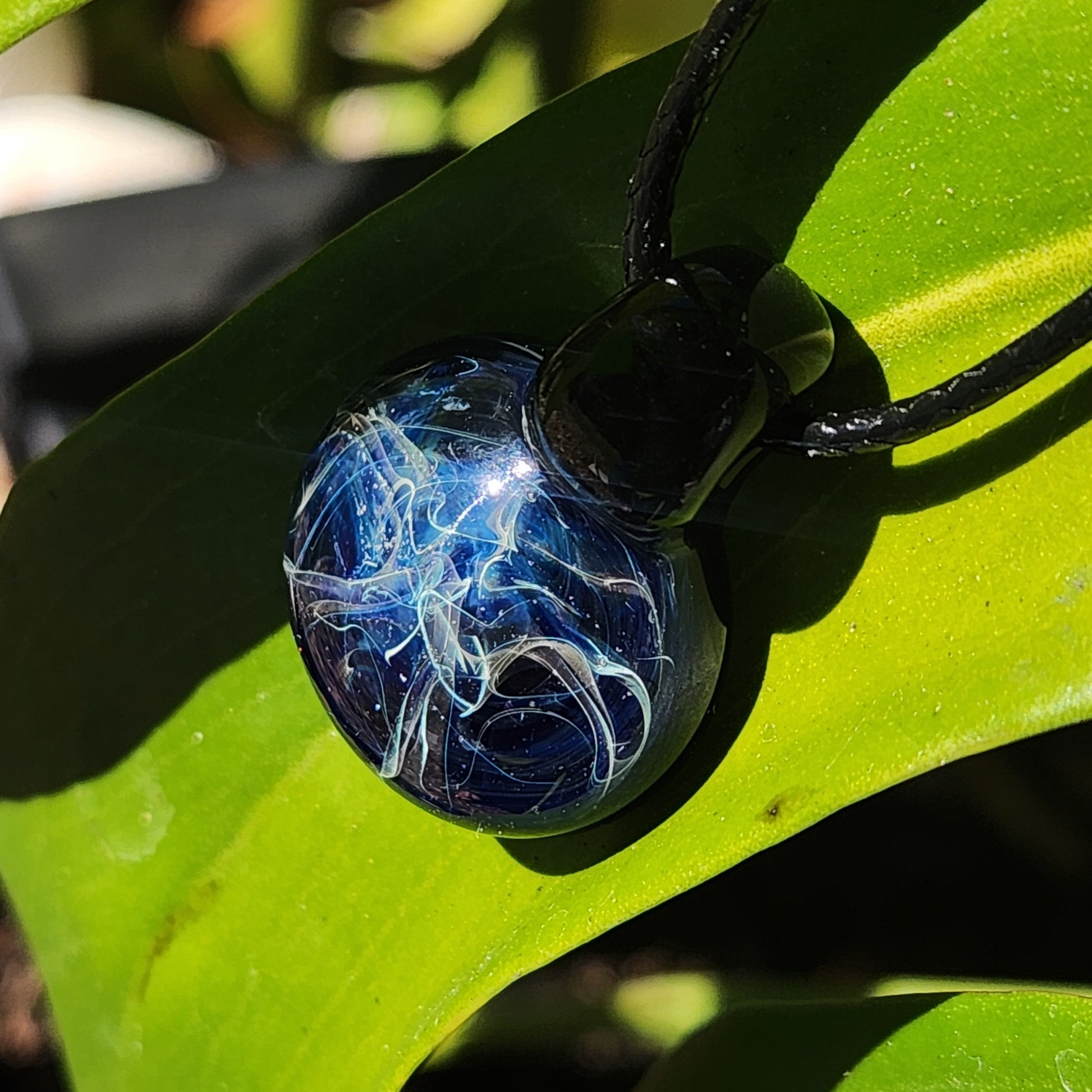Unique birthday gift pendant featuring a small blue swirl glass charm, displayed on a green leaf in natural light.