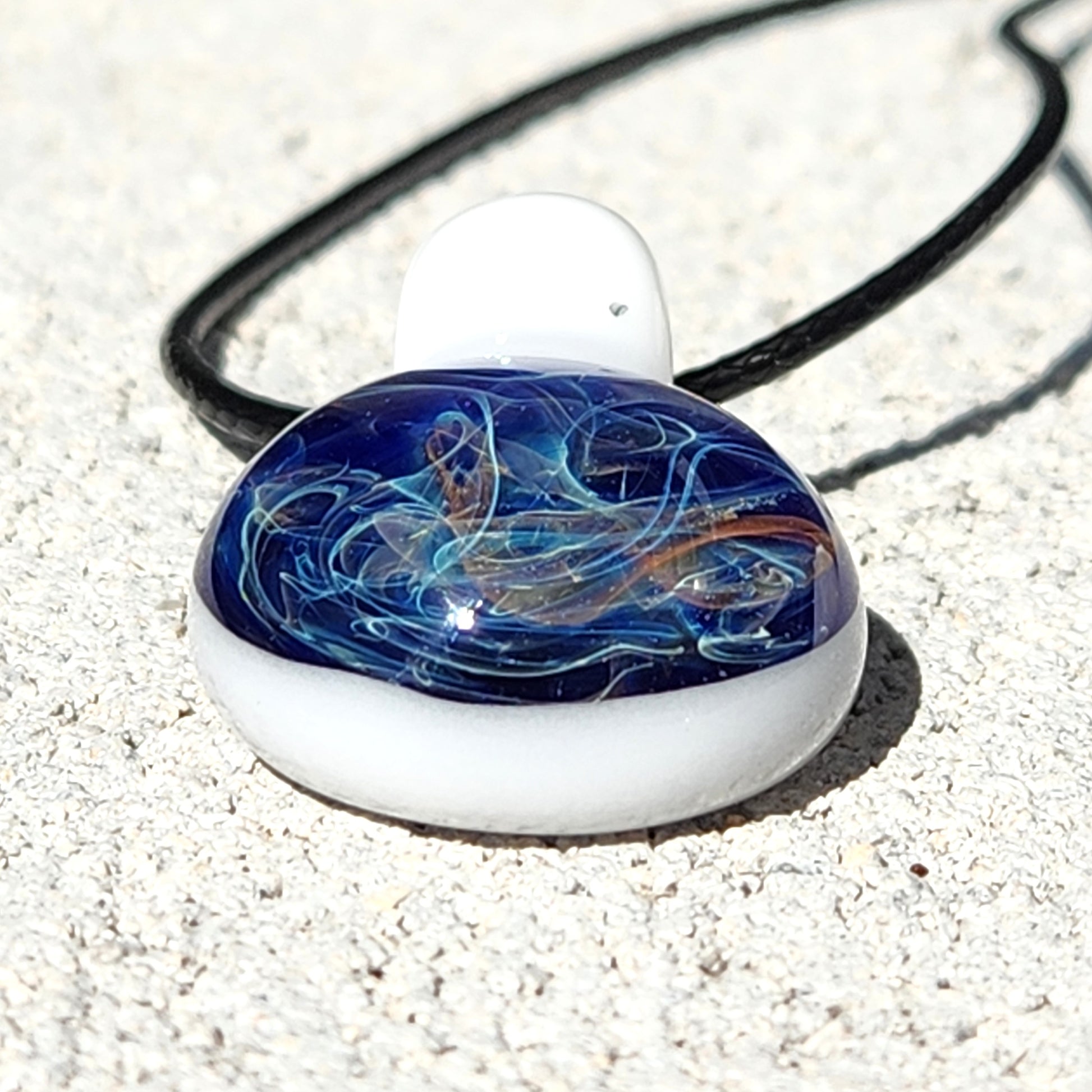 Flat-lay of Deep Blue Galaxy Pendant Necklace with shimmering blue, gold, and white swirls, handmade glass charm on sand.