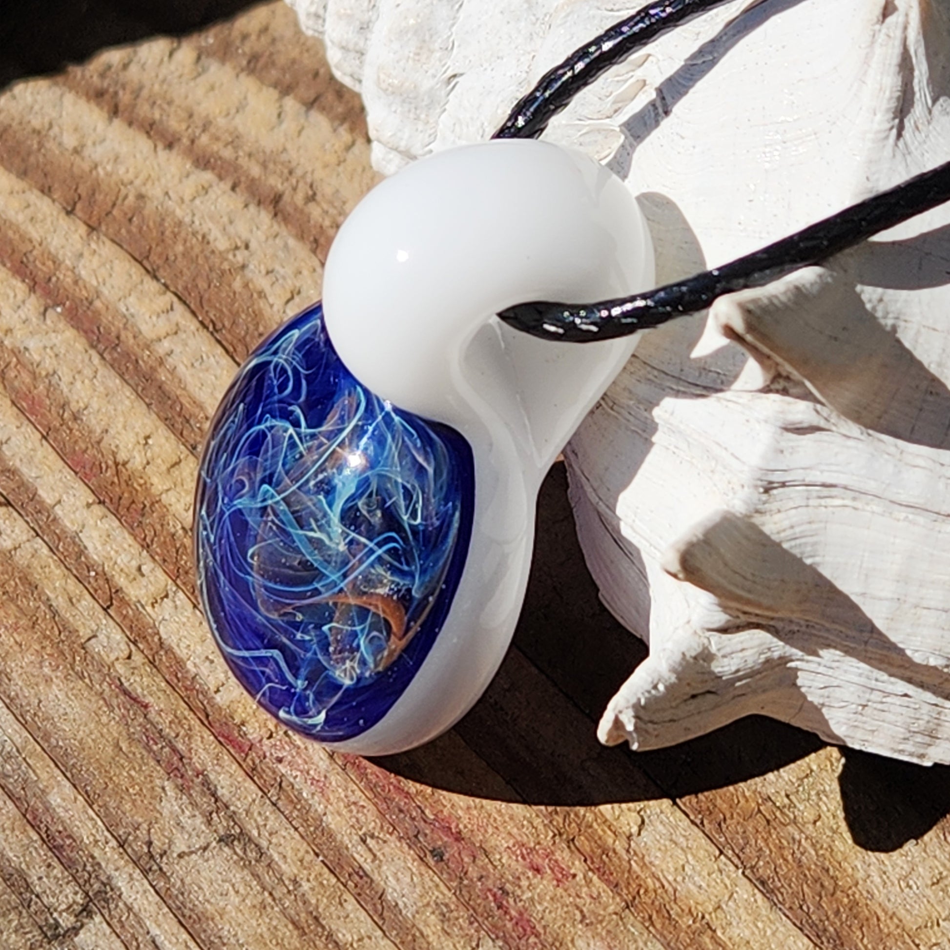 Deep Blue Galaxy Pendant Necklace with swirling galaxy design, handmade glass jewelry resting on a wooden surface near a shell.