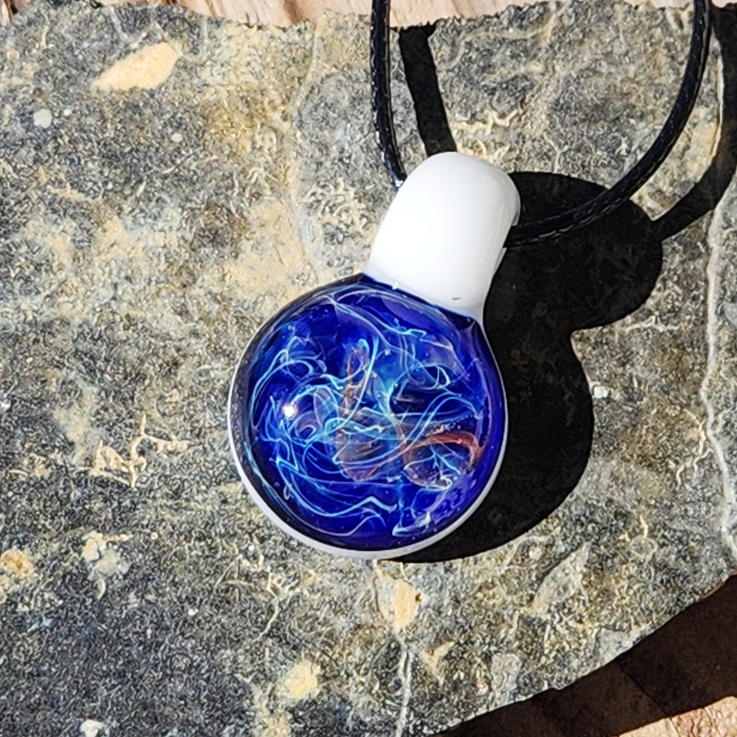 Unique glass pendant necklace featuring a deep blue galaxy design with white and gold swirls, resting on a textured stone surface.