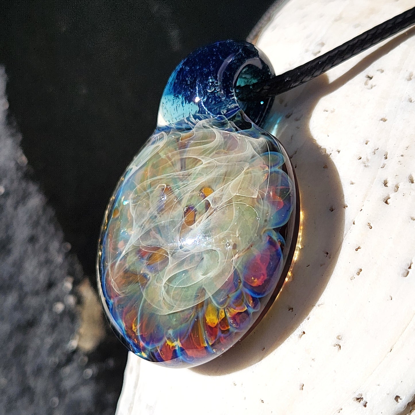 Angled view of handmade glass pendant necklace with deep blue top and colorful swirling center, resting on a white seashell.