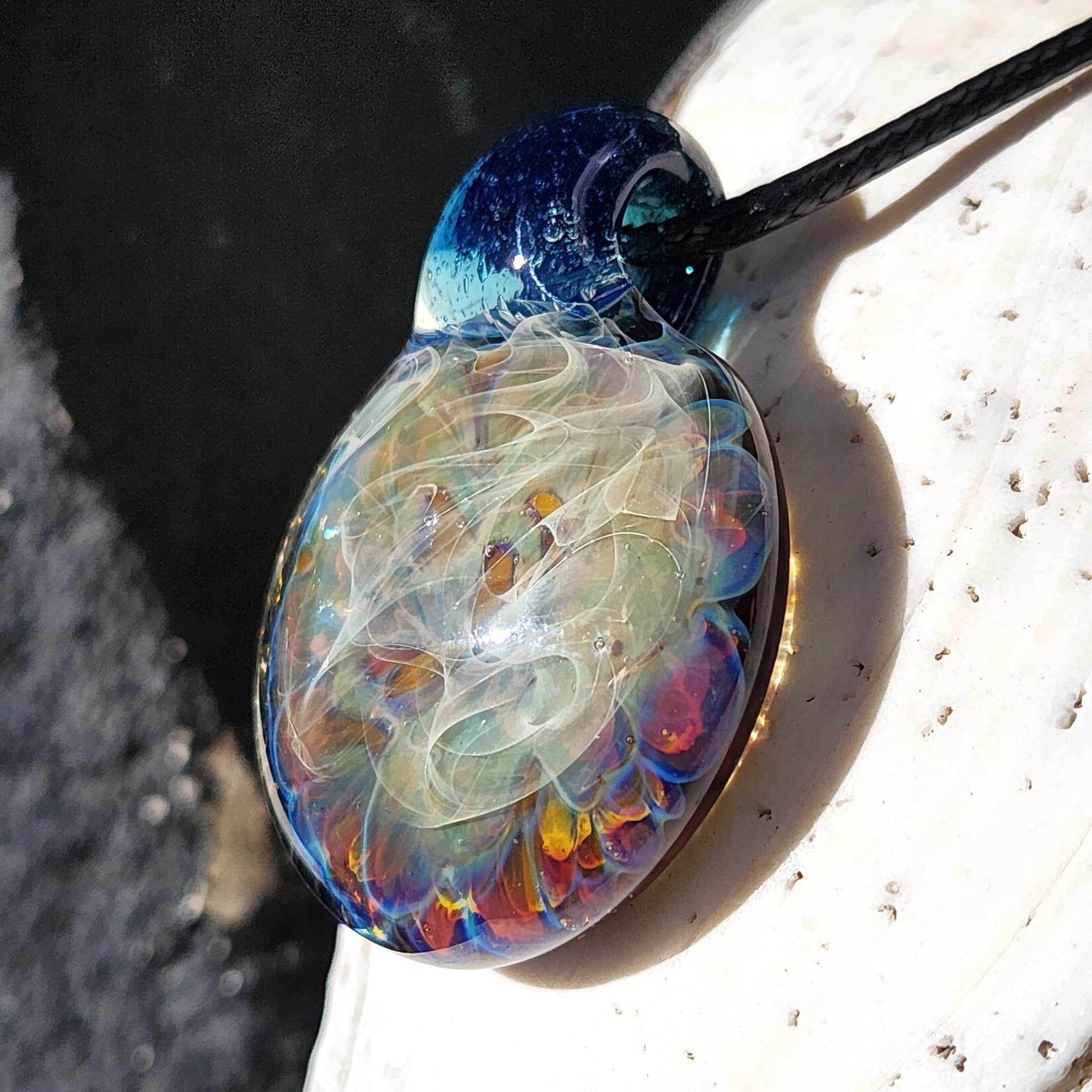 Angled view of handmade glass pendant necklace with deep blue top and colorful swirling center, resting on a white seashell.