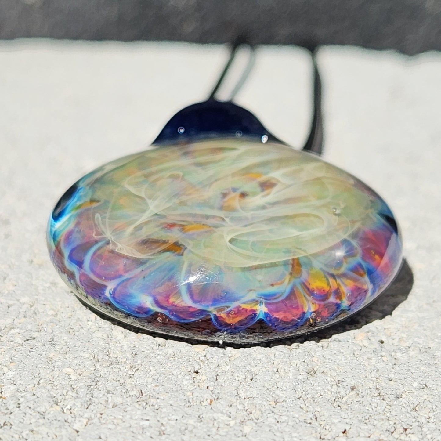 Close-up of handmade blown glass pendant necklace showing vibrant rainbow hues and layered glass texture on a light stone surface.