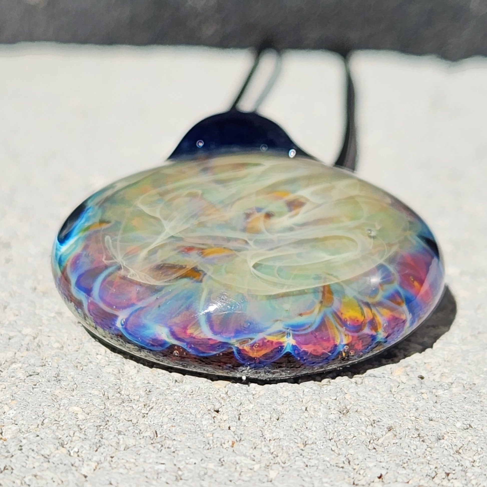 Close-up of handmade blown glass pendant necklace showing vibrant rainbow hues and layered glass texture on a light stone surface.