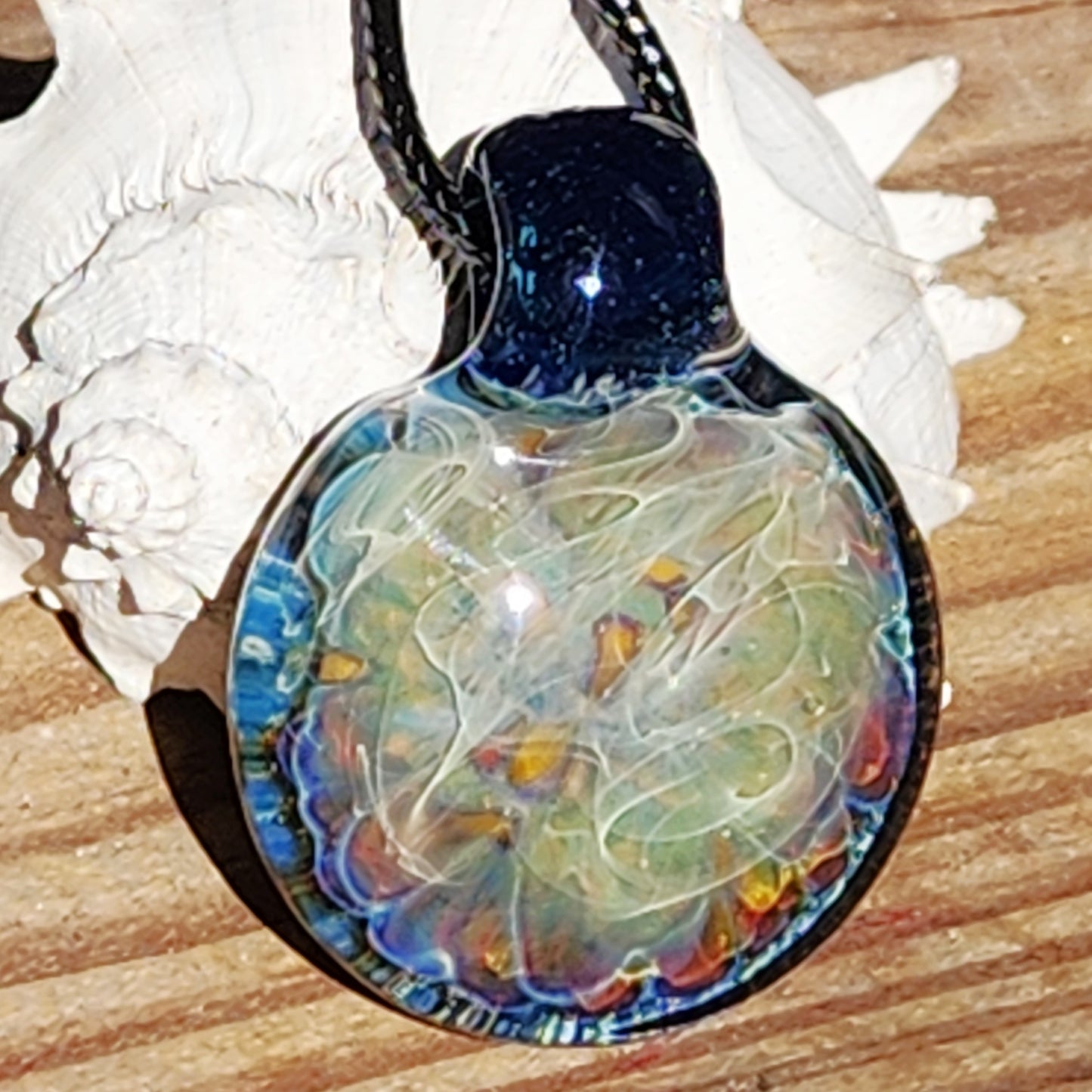 Handmade blown glass pendant necklace with swirling blue, silver, and rainbow tones, displayed on wood with seashells for a natural jewelry look.