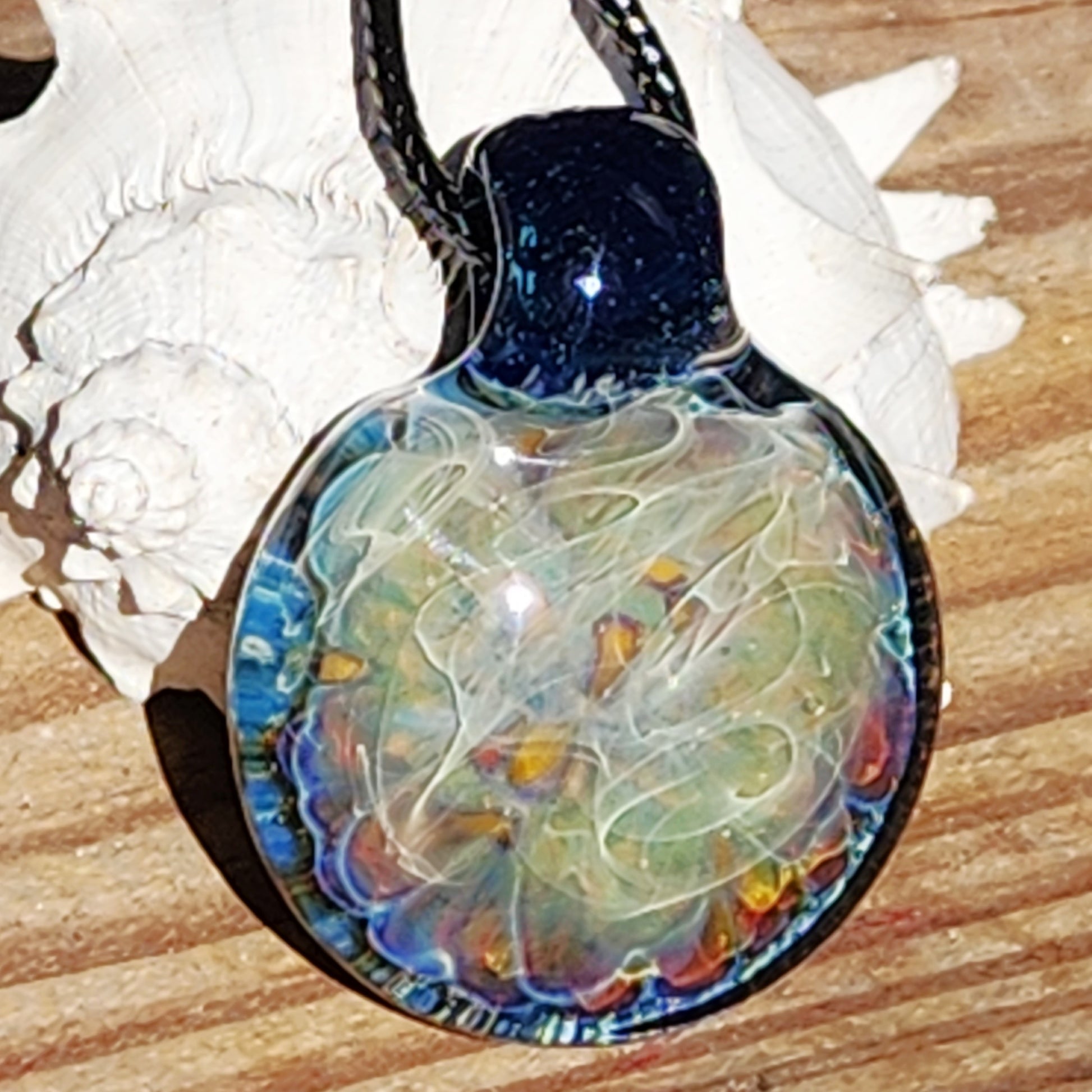 Handmade blown glass pendant necklace with swirling blue, silver, and rainbow tones, displayed on wood with seashells for a natural jewelry look.