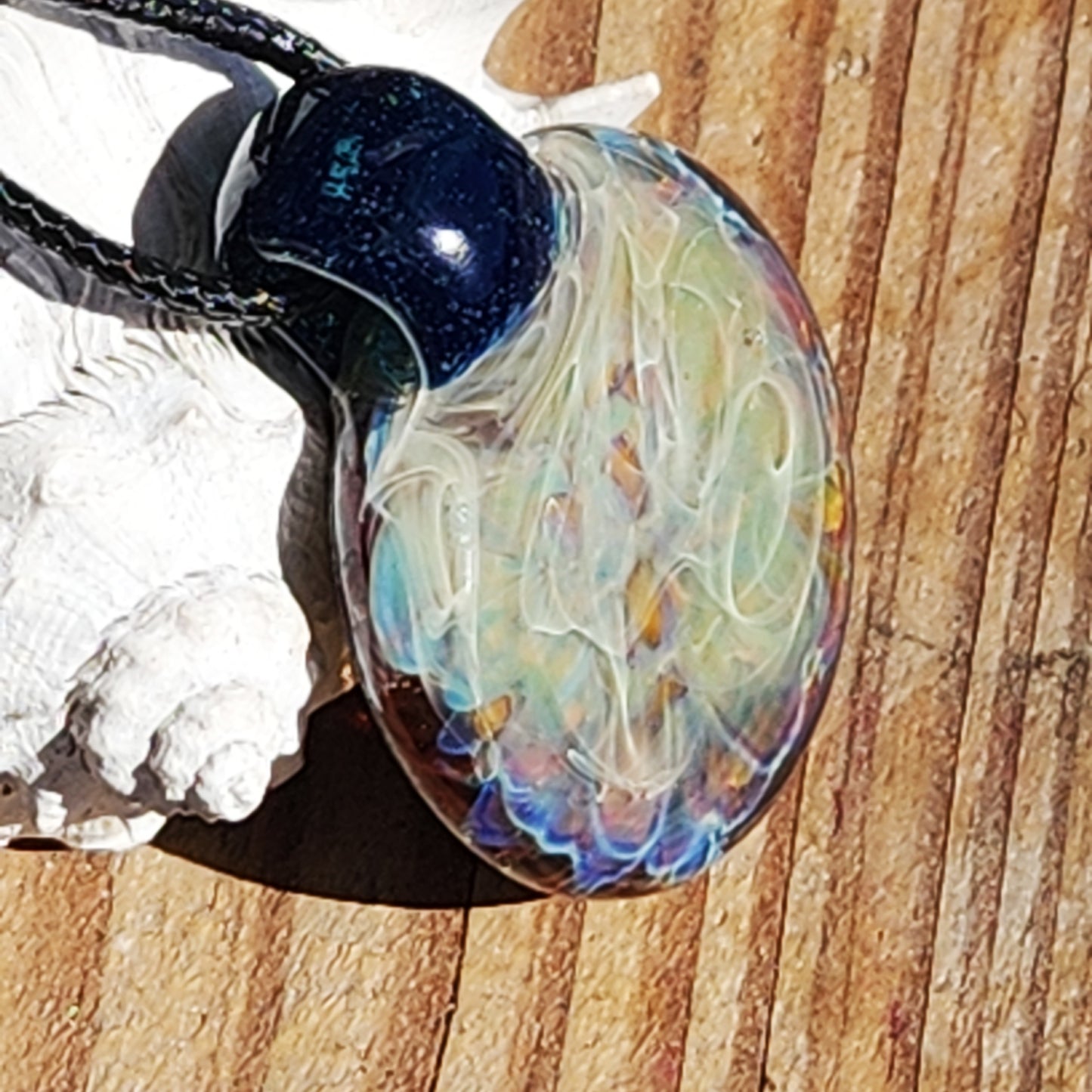 Artistic handmade glass pendant necklace with silver fuming and colorful layers, displayed on wood with natural shell accents.