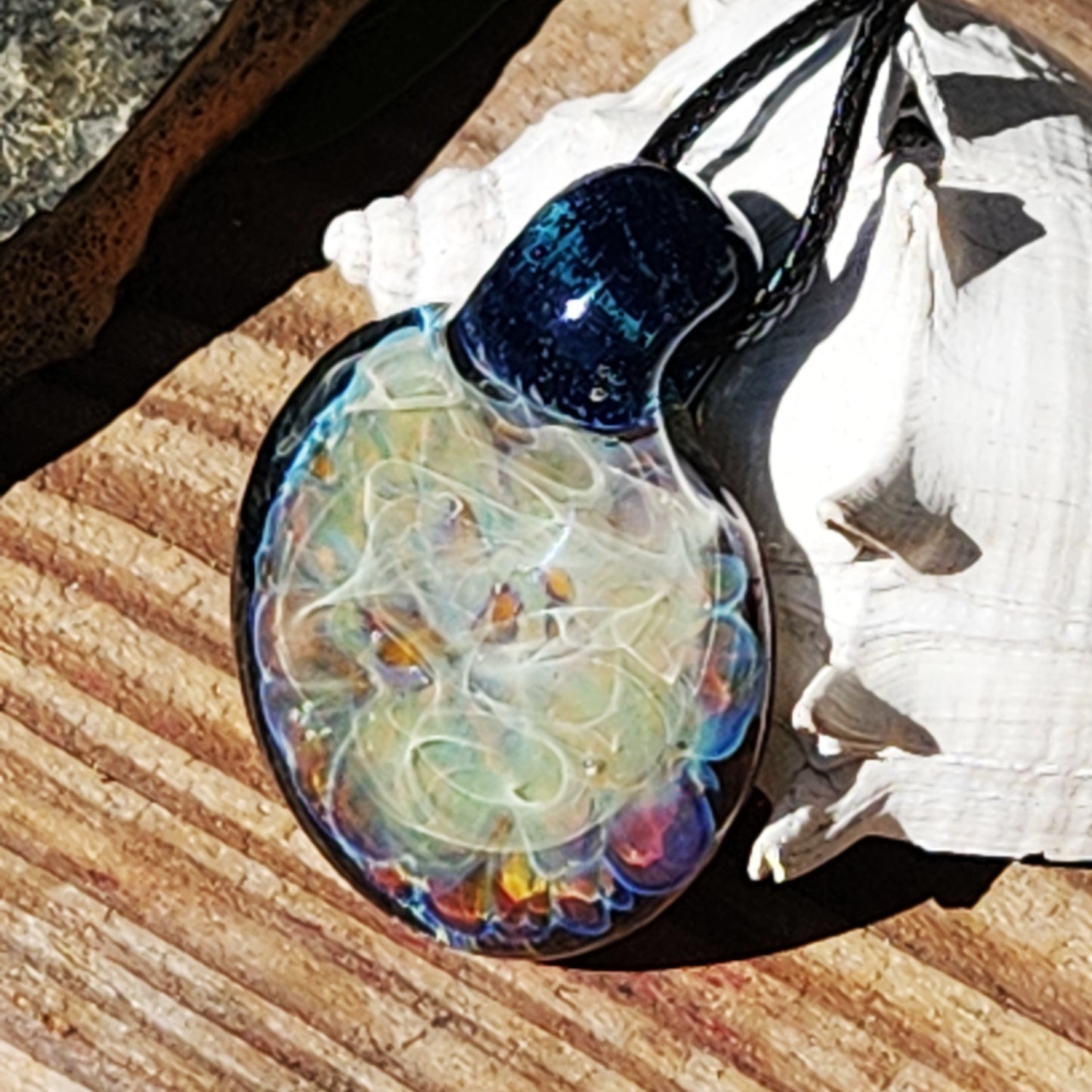 Colorful handmade glass pendant necklace with glowing swirls and silver accents, placed on rustic wood with seashell decor.