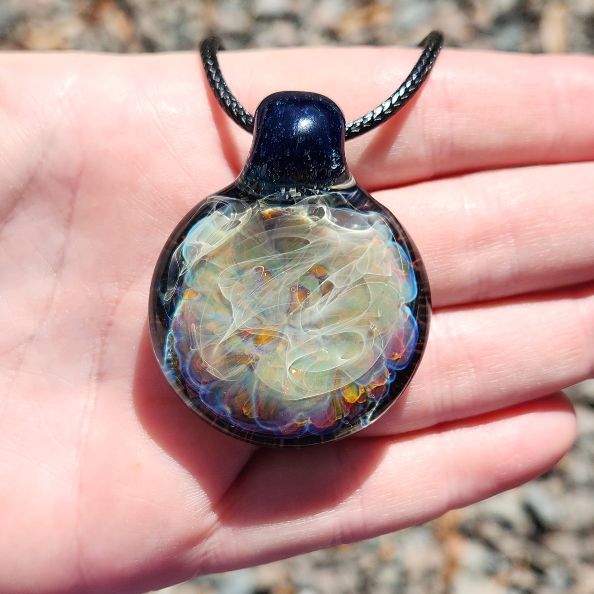 Hand holding handmade blown glass pendant necklace, highlighting its size and intricate swirling design in natural sunlight.