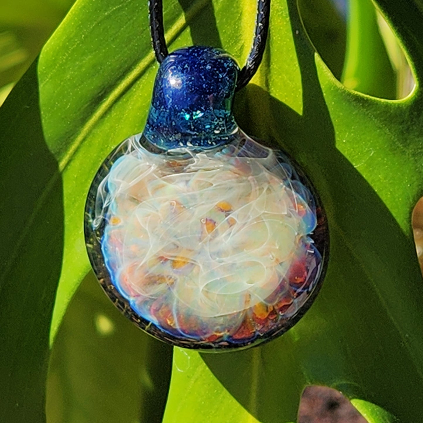 Handmade blown glass pendant necklace hanging against green foliage, showcasing its glowing, multicolor design and silver fumed details.