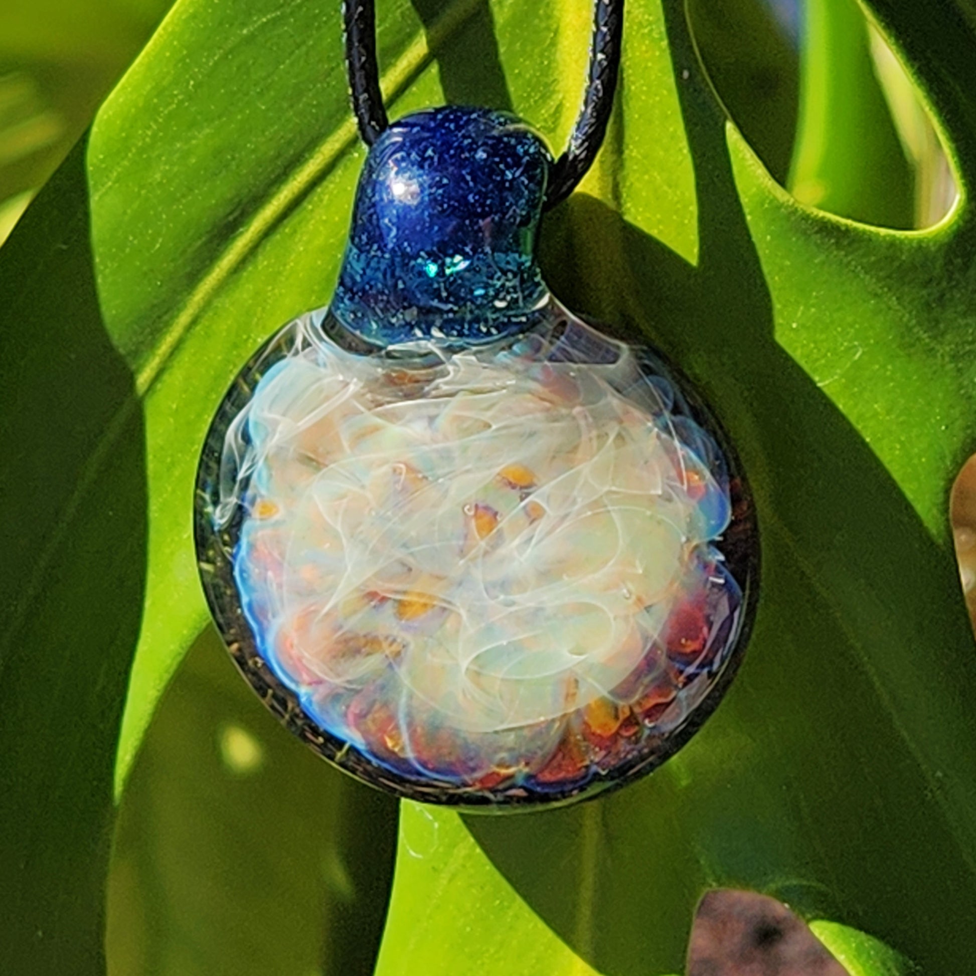 Handmade blown glass pendant necklace hanging against green foliage, showcasing its glowing, multicolor design and silver fumed details.
