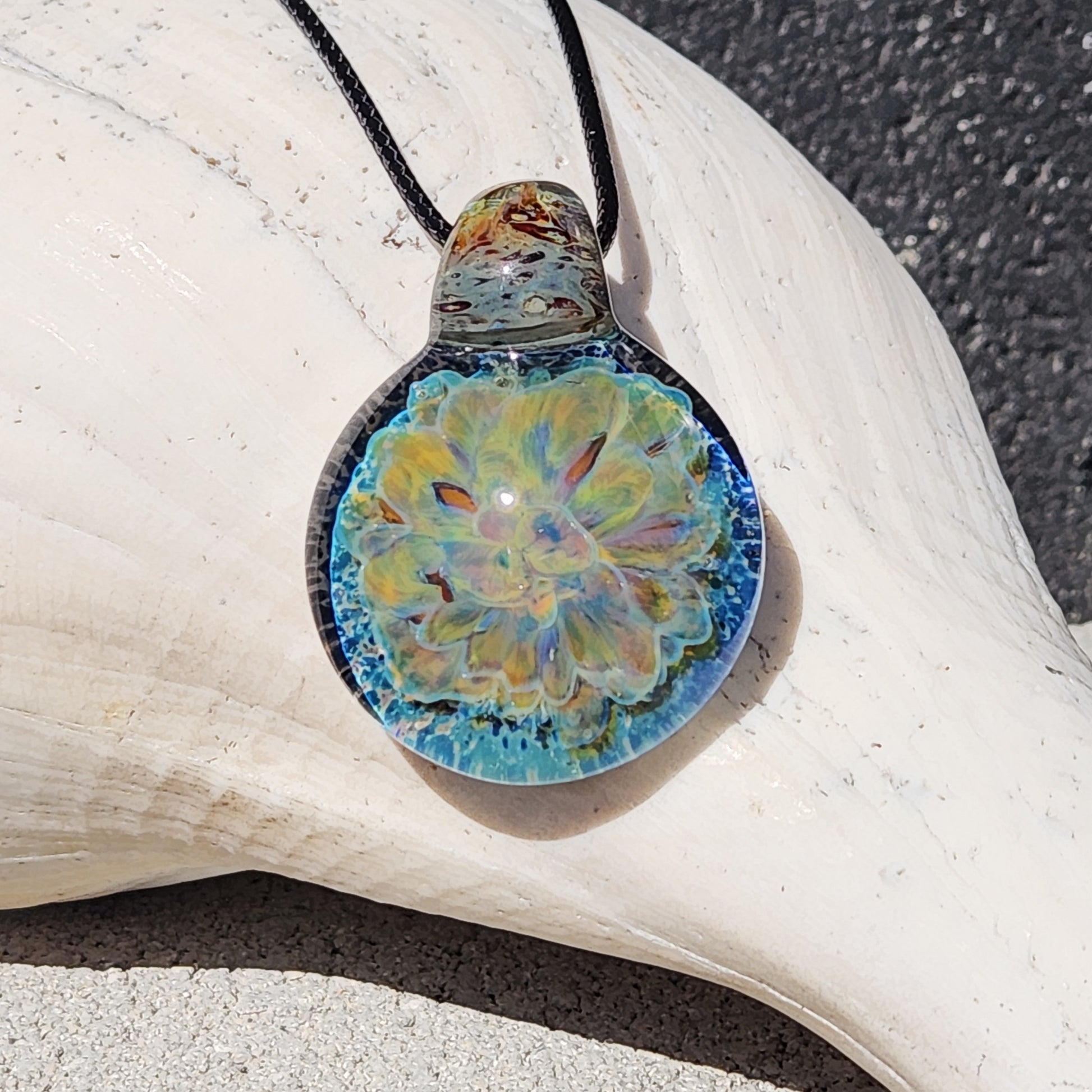 Handmade glass pendant necklace featuring a glowing yellow and blue floral design, displayed on a white seashell background.