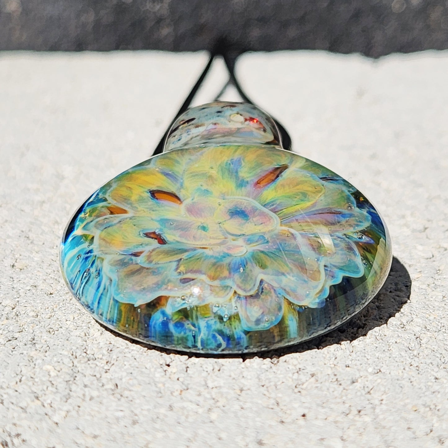 Front-facing view of the handmade glass pendant necklace with a radiant yellow-blue flower design, lying on a light stone surface.