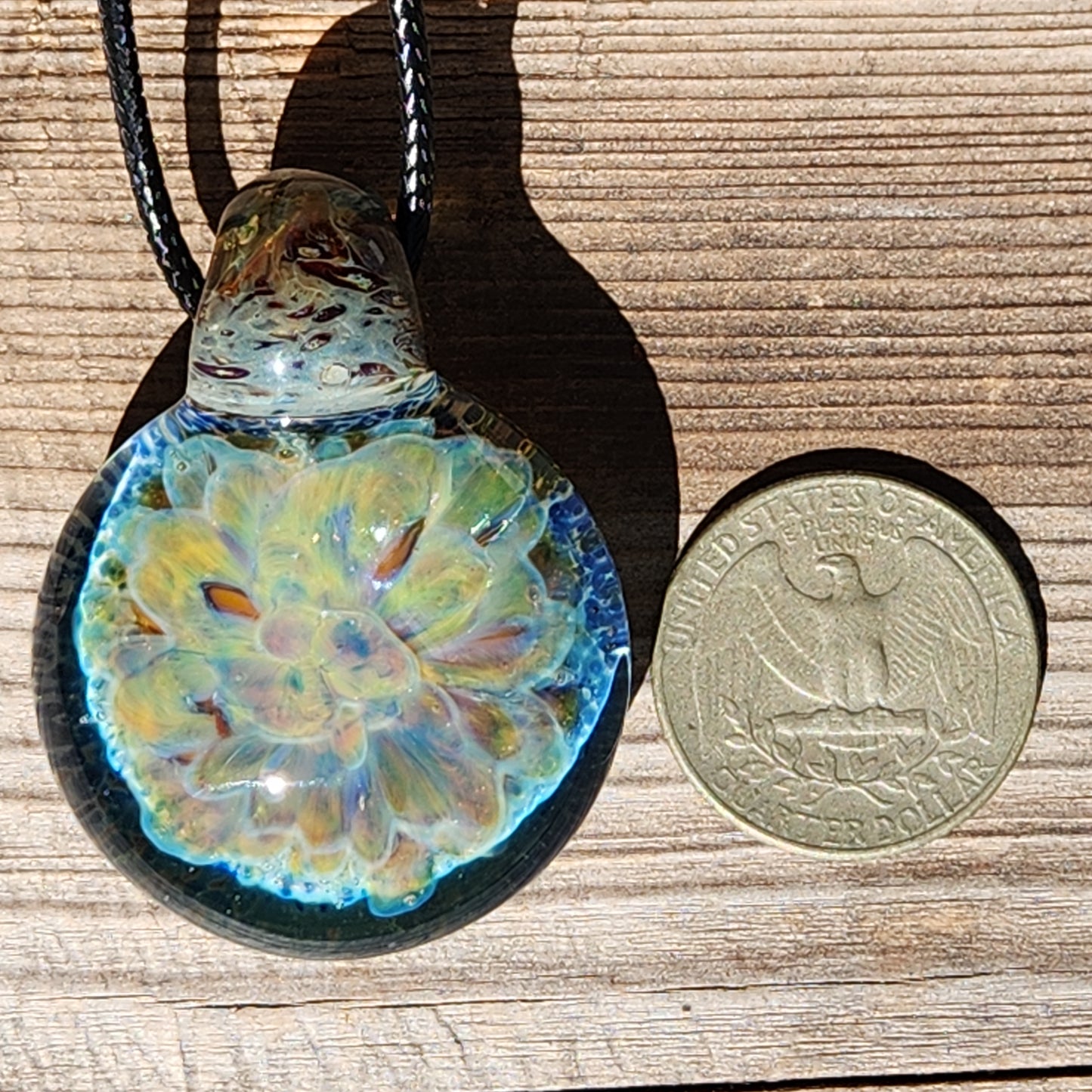 Handmade glass pendant necklace next to a US quarter for size comparison, featuring a glowing yellow and blue floral design.