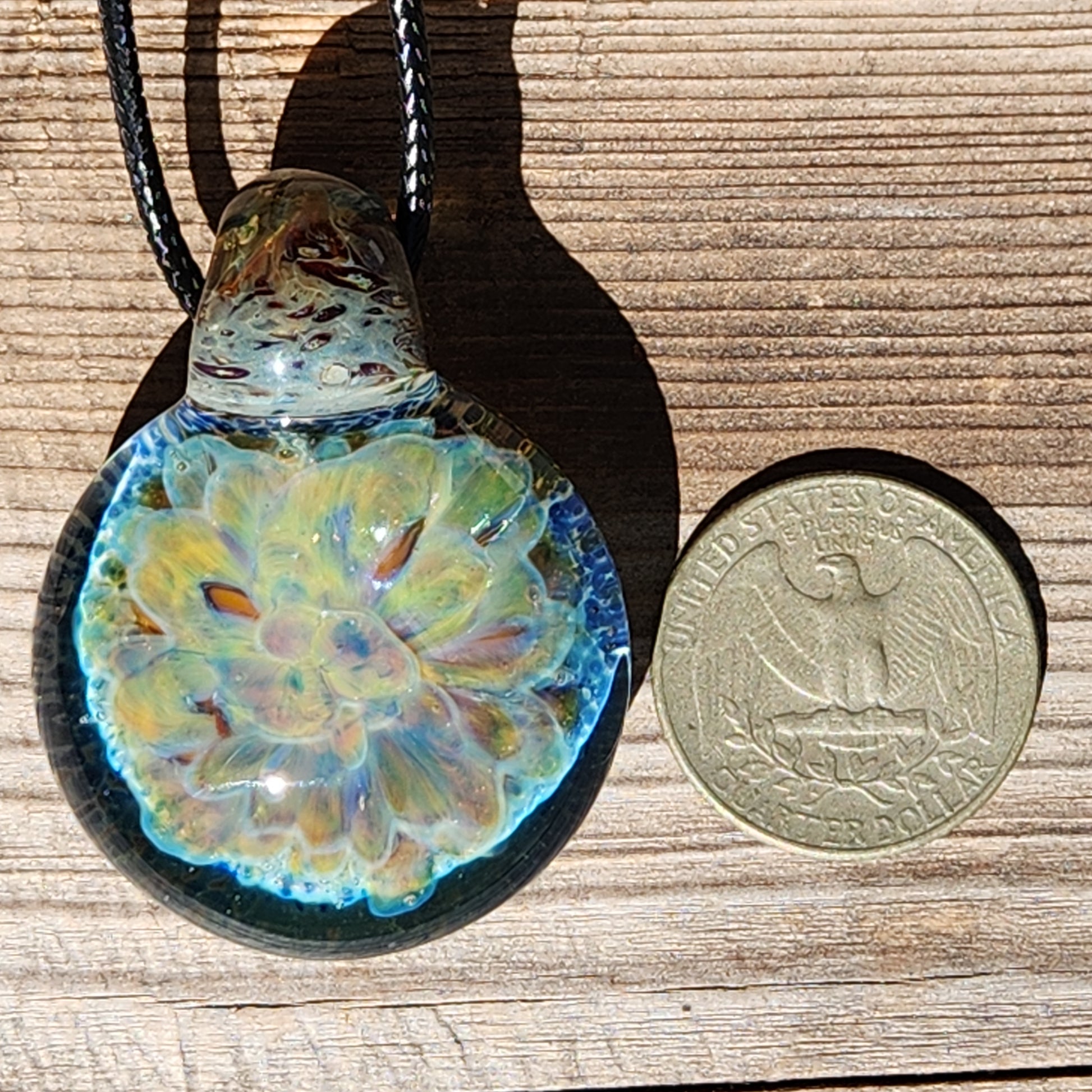 Handmade glass pendant necklace next to a US quarter for size comparison, featuring a glowing yellow and blue floral design.