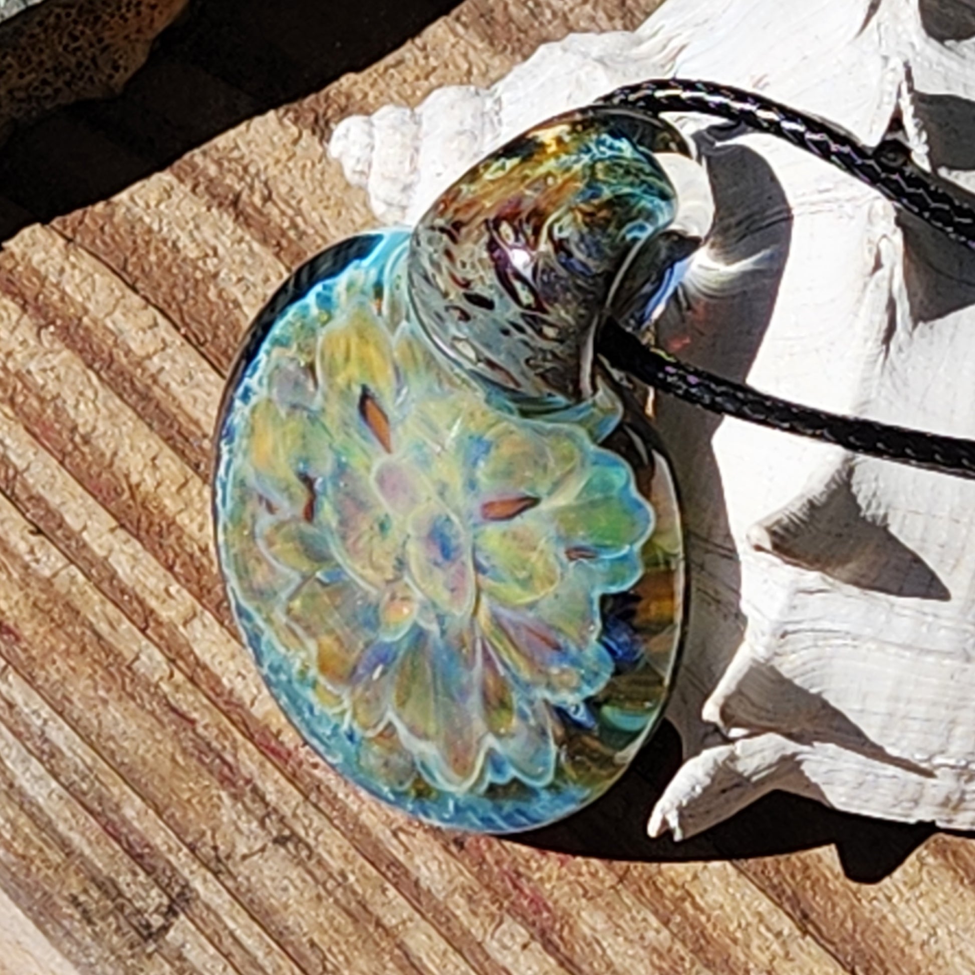 Artistic handmade glass pendant necklace with a vivid yellow and blue flower design, displayed on a wooden surface with seashell.