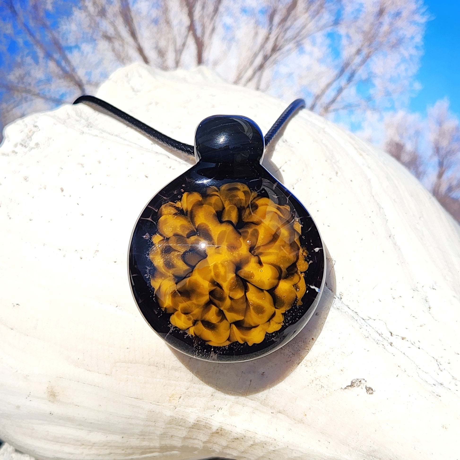 Handmade glass pendant necklace with bold yellow and black spiral design, displayed on a white seashell with a blue sky background.