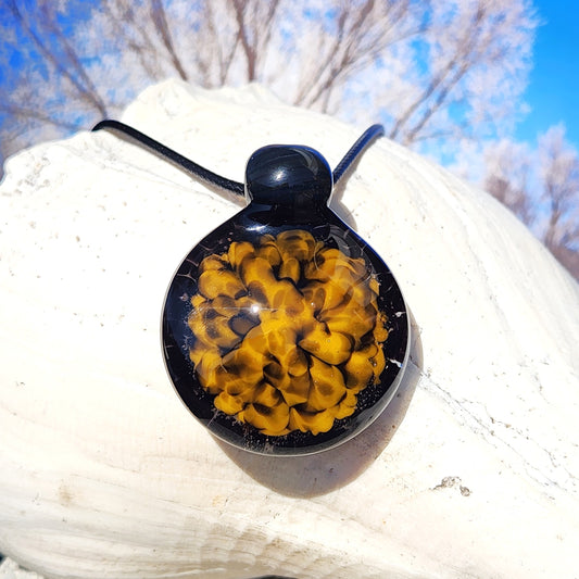 Handmade glass pendant necklace with bold yellow and black spiral design, displayed on a white seashell with a blue sky background.