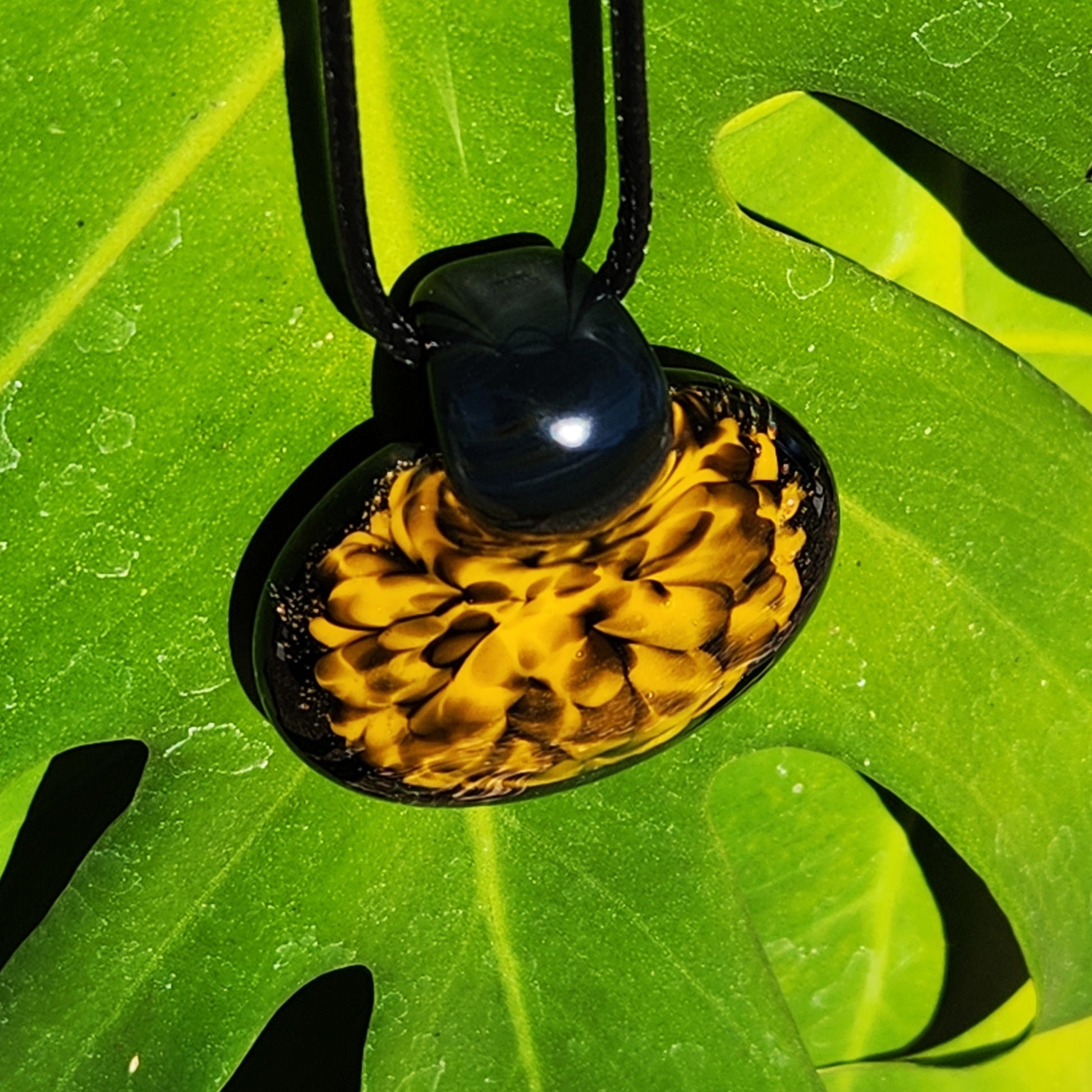 Angled view of handmade glass pendant necklace with bold yellow spiral pattern, hanging on a green leaf in sunlight.