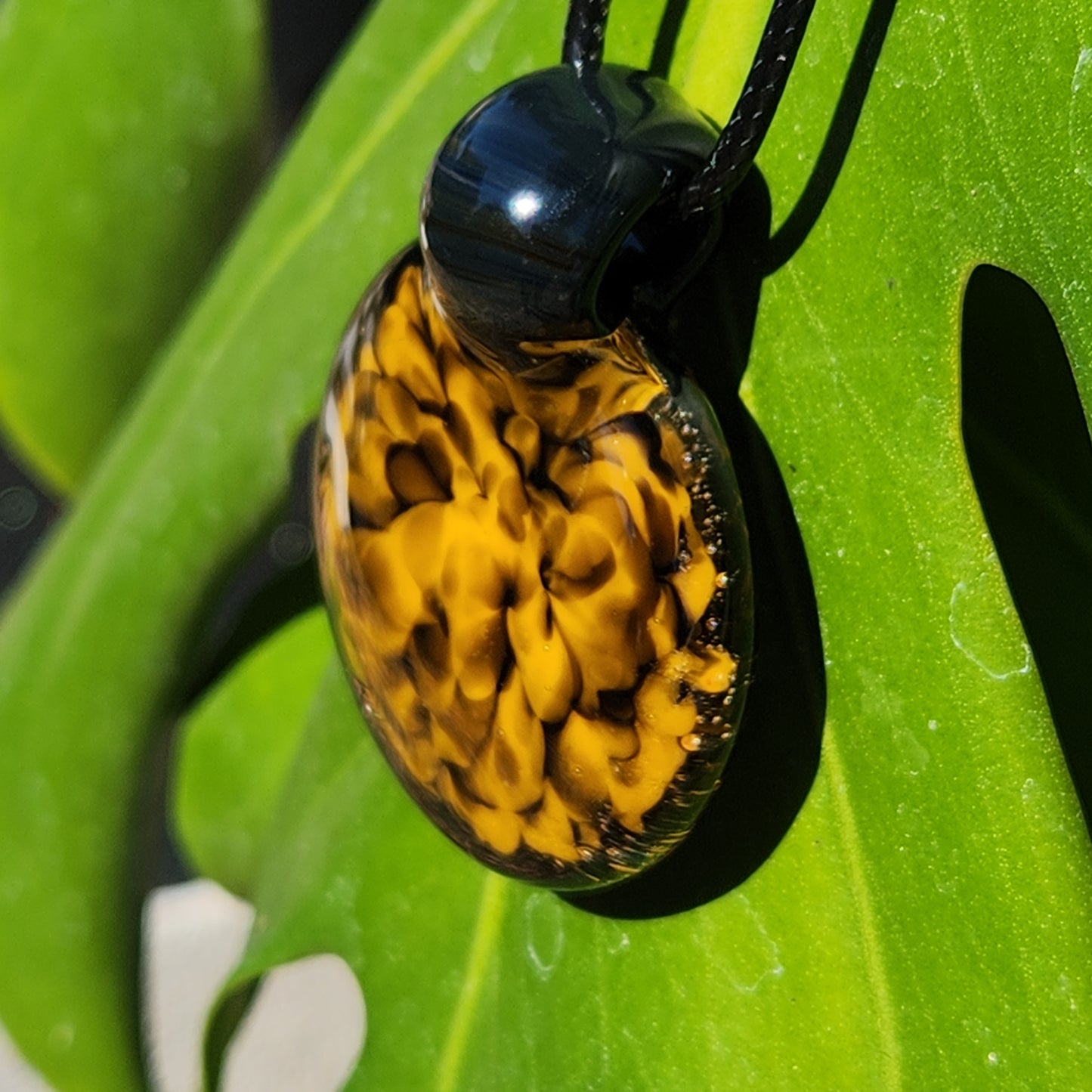 Angled view of handmade yellow and black spiral glass pendant resting on a green leaf, showcasing its curved shape.
