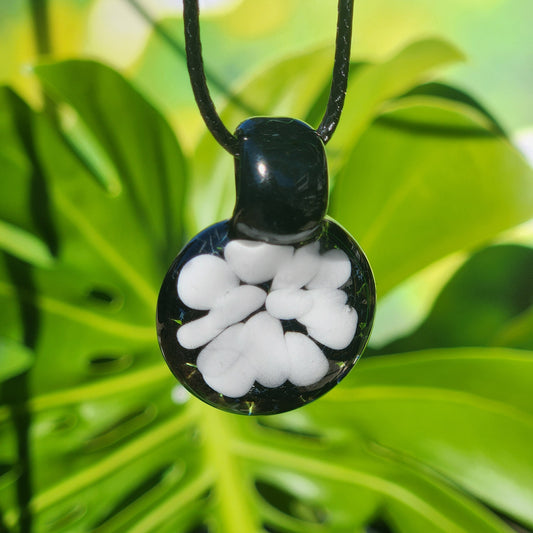 Minimalist white glass pendant with floral design on black borosilicate base, handmade in the USA, displayed on green leaf background.