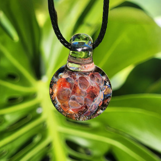 Handmade unique glass pendant with red and multicolor crushed glass design, hanging on black cord, shown against green leaf background.