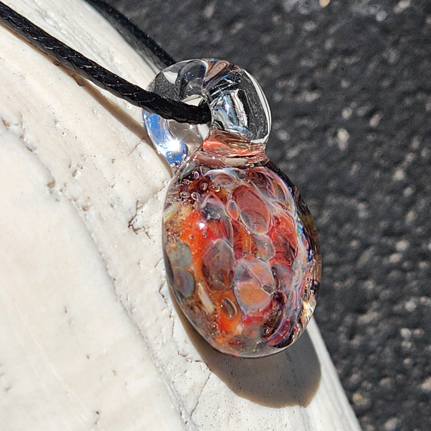 Angled view of small handmade glass pendant with fiery red and orange crushed glass, lying on a white shell.