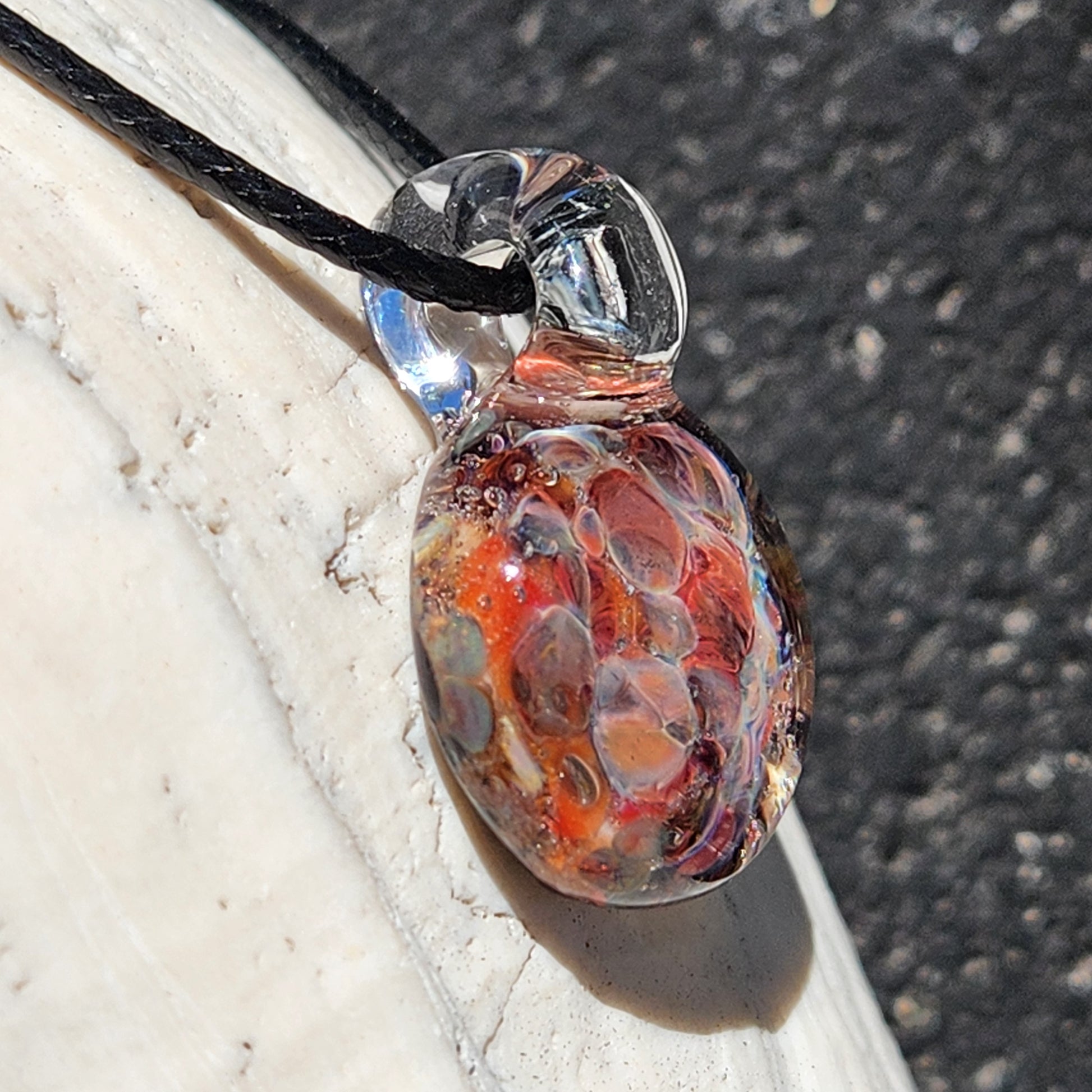Angled view of small handmade glass pendant with fiery red and orange crushed glass, lying on a white shell.