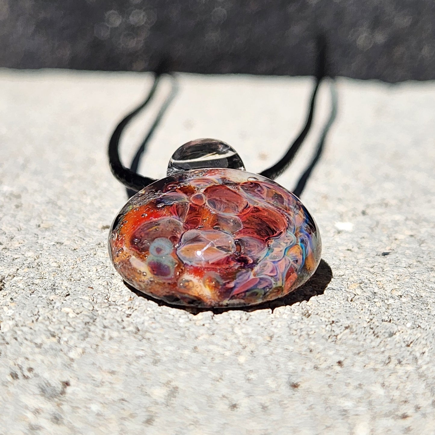 Front view of handmade glass pendant with vibrant red and purple bubble design, placed on concrete with black cord.