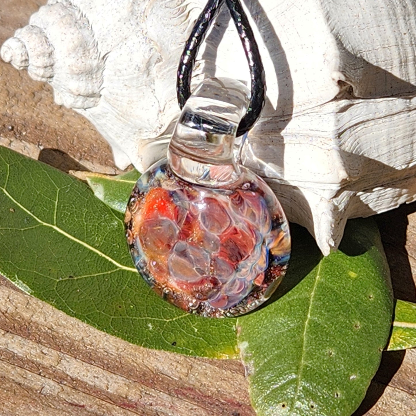 Handmade borosilicate glass pendant resting on green leaves and wood, featuring a vibrant red and orange crushed glass core.