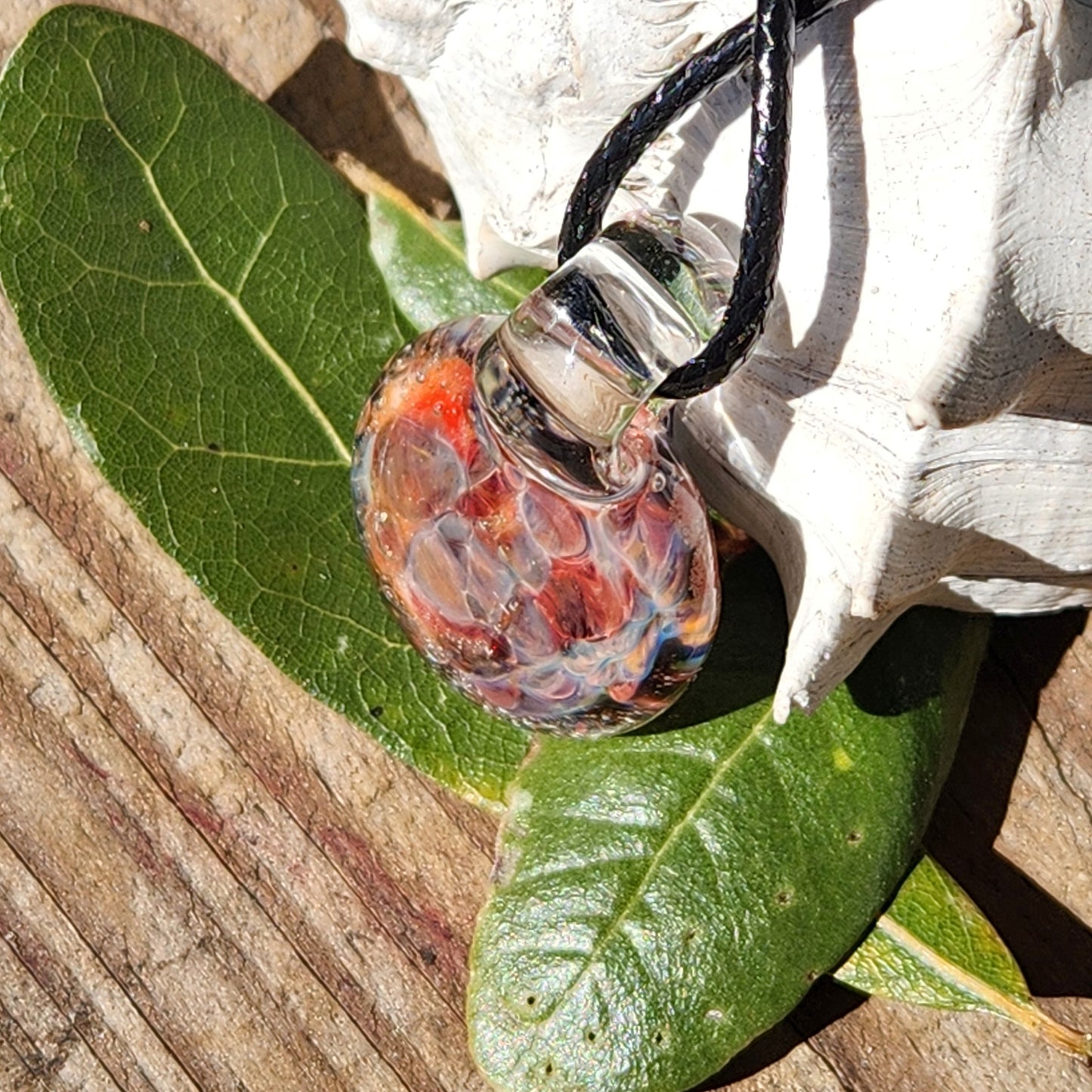 Angled view of small glass pendant with red and purple crushed glass, displayed on green leaves and wood.